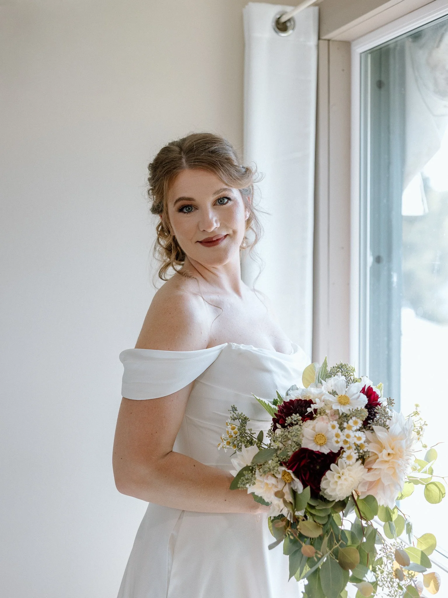 A bride with curly blonde hair in a white off-shoulder wedding gown holding a large bouquet of white, red, and pink flowers and greenery, standing near a window.