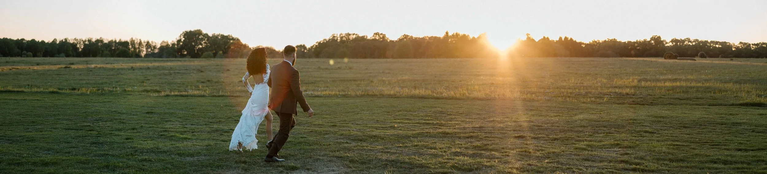 A couple dressed in wedding attire walking across a grassy field during sunset.
