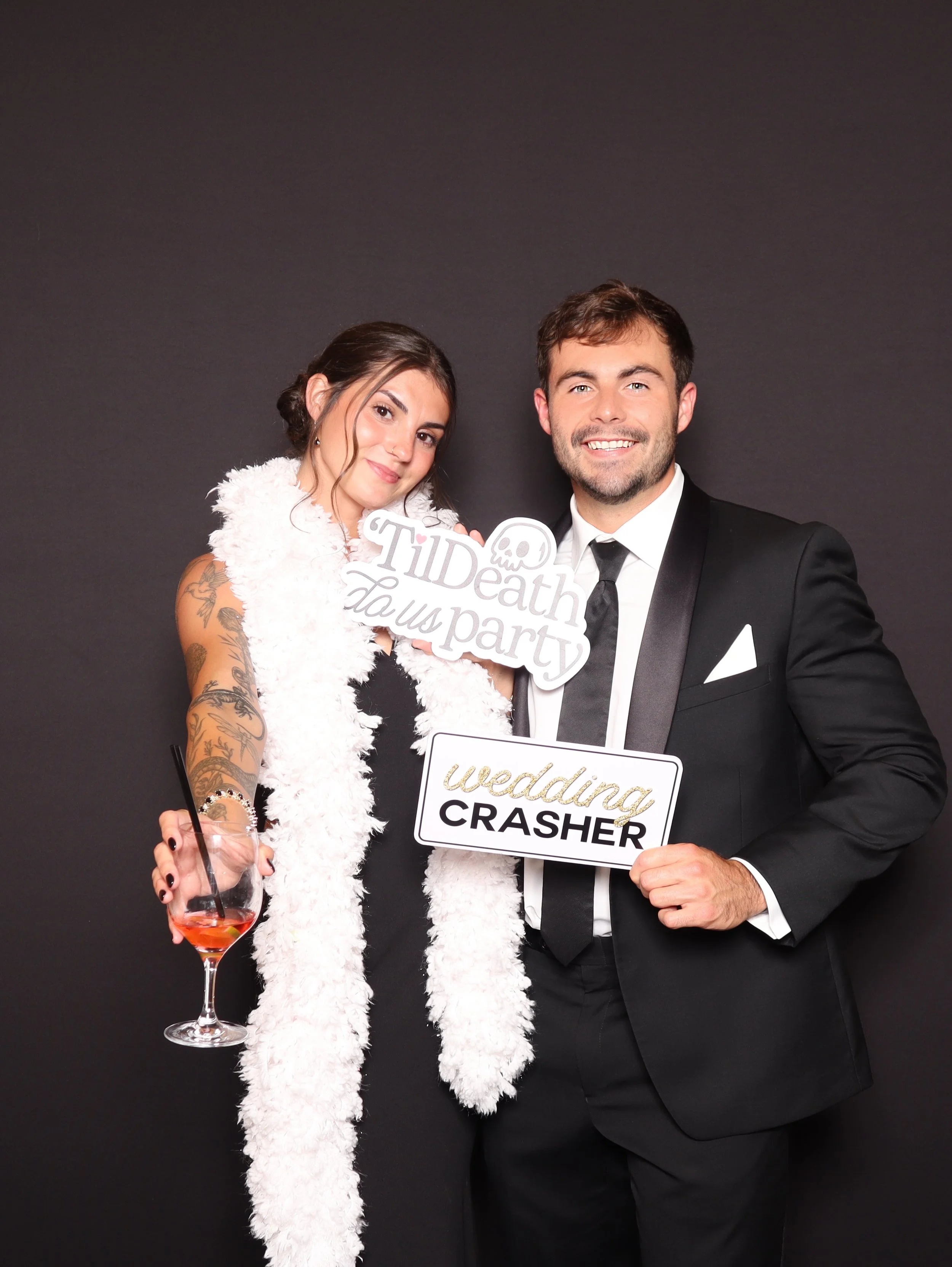 A bride and groom at a wedding reception holding signs, with the bride holding a drink, standing against a black background.