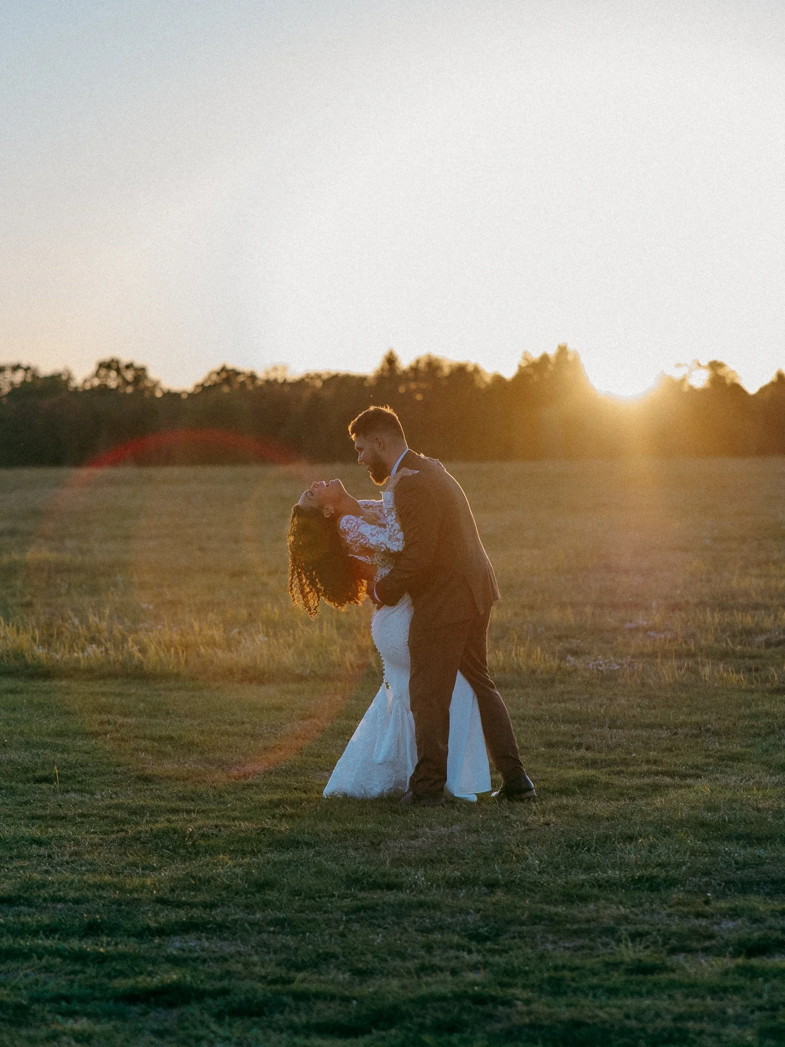 A newlywed couple dancing outdoors in a field at sunset, with the groom dipping the bride.
