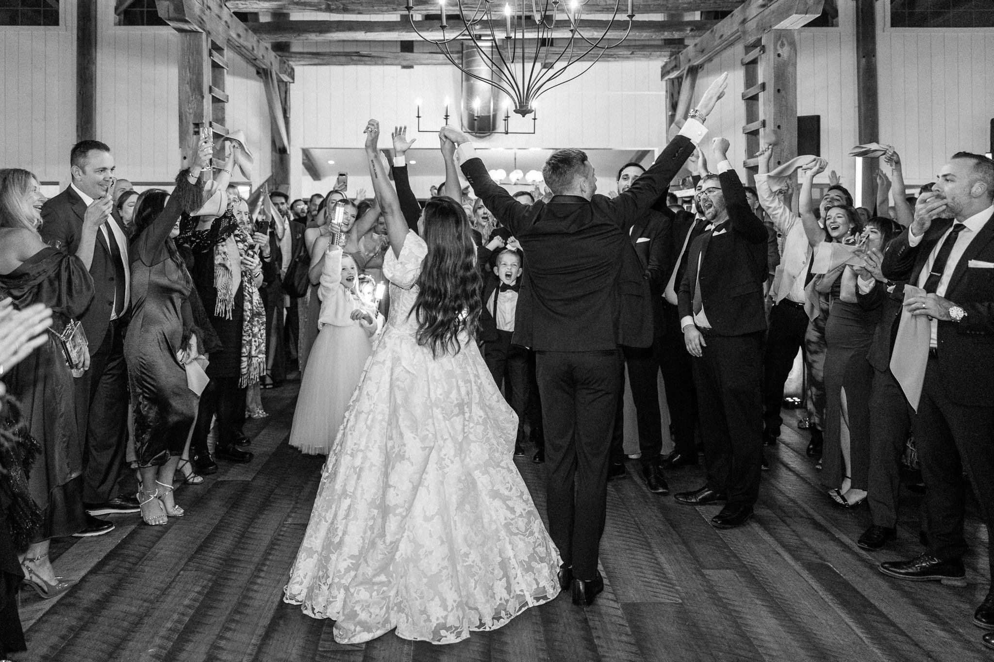 A bride and groom dancing at their wedding reception surrounded by guests who are smiling, raising their hands, and holding drinks in a rustic-style venue with wooden beams and chandeliers.