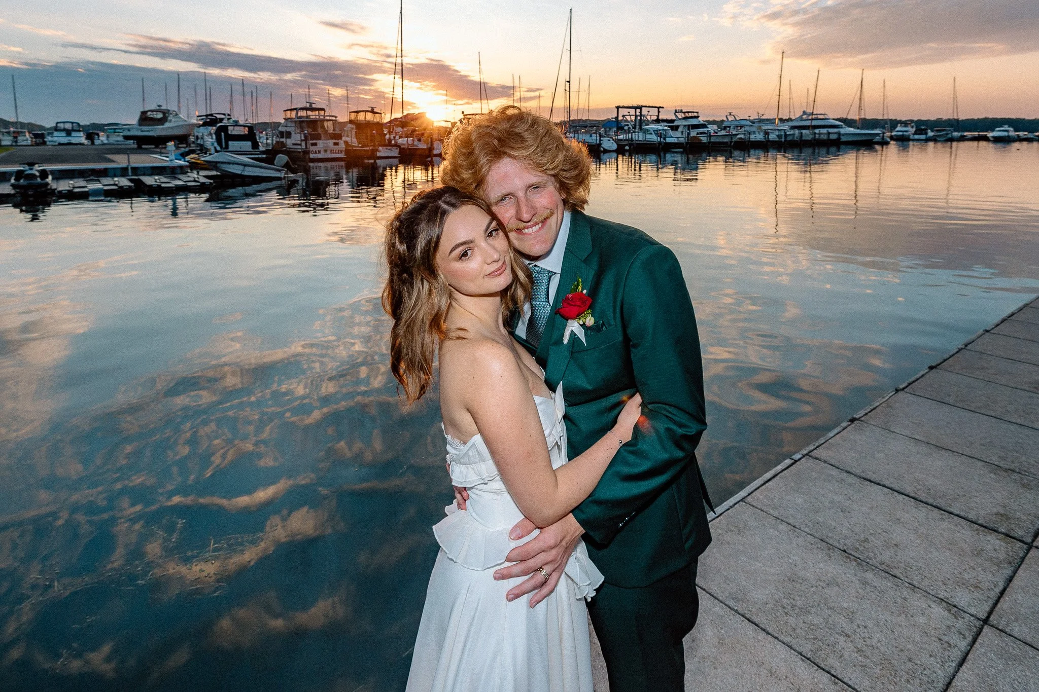 bride and groom at their wedding hug each other and look at the viewer with the Erie Yacht club and Lake Erie in the background as the clouds reflect of the water and the sun is setting.