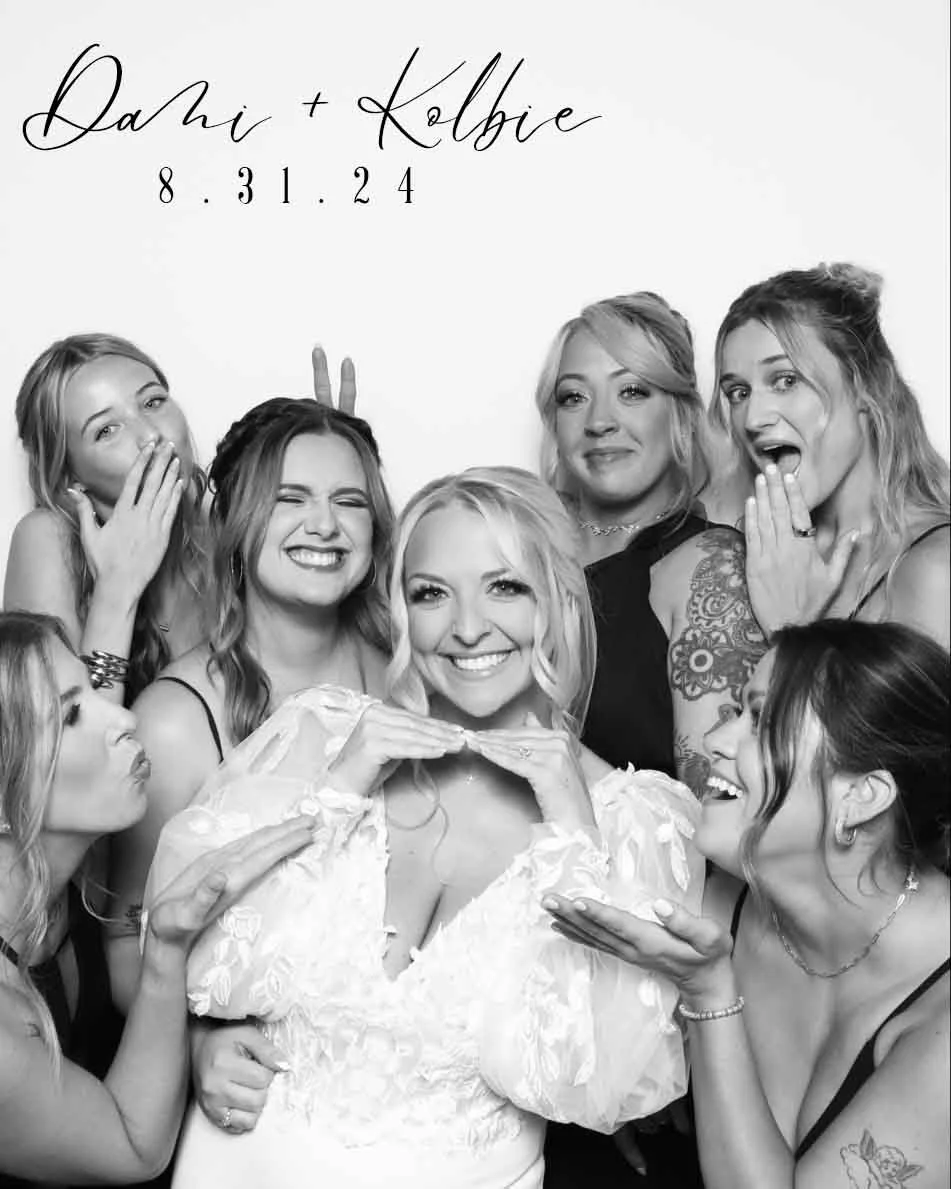 Black and white photo of seven women, one in a white dress, celebrating a wedding. The woman in the white dress is in the center, smiling, with her hands under her chin. The other women are surrounding her, making playful expressions and gestures. Te