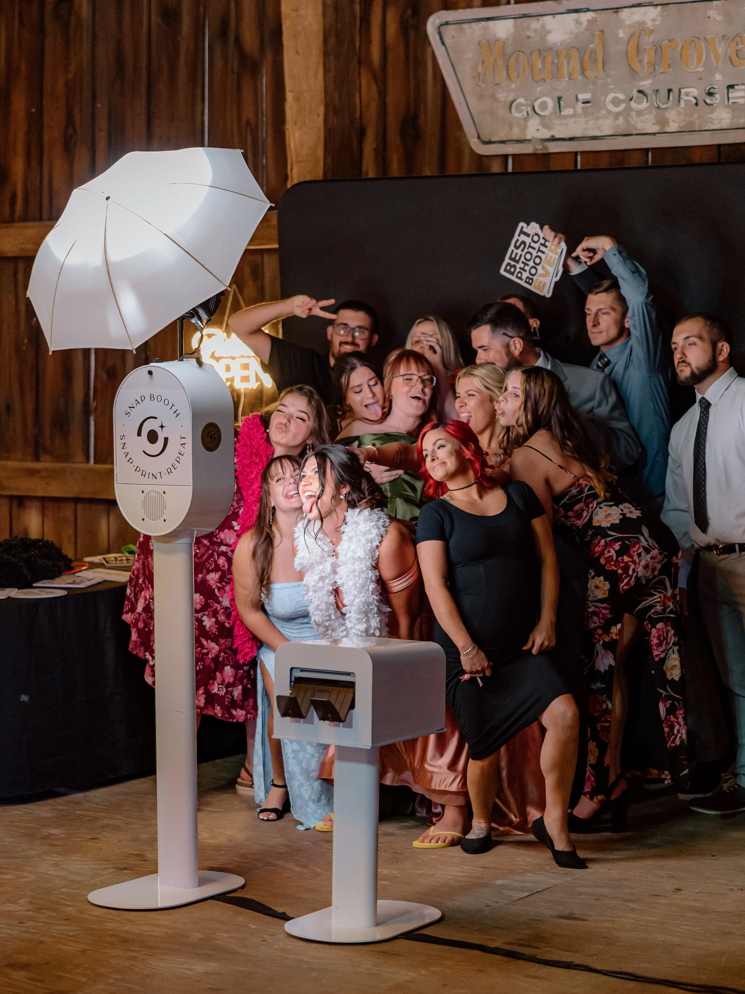 Group of people posing for a photo in a photo booth area with a large umbrella light, a photo booth printer, and a sign that says 'SNAP PRINT REPEAT.' Everyone is smiling and making funny faces.