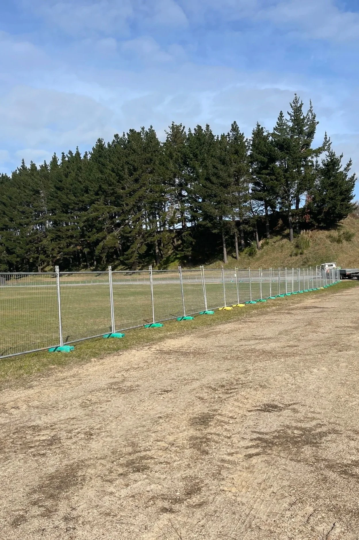 A row of green trees behind a metal fence on a grassy area, with a truck parked near the trees under a blue sky with some clouds.