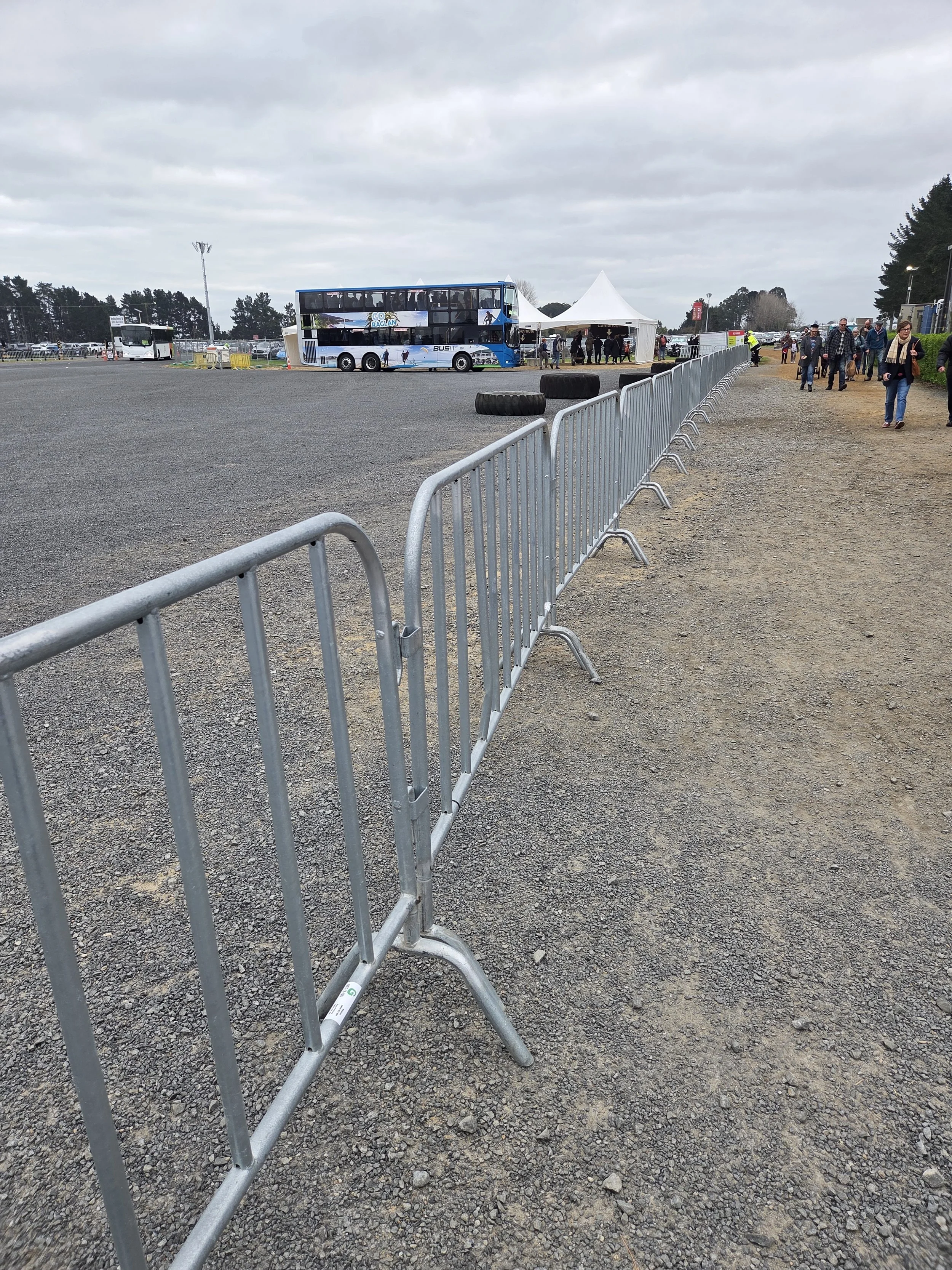 People walking past metal barricades at an outdoor event with a large blue double-decker bus and a white tent in the background, under cloudy skies.