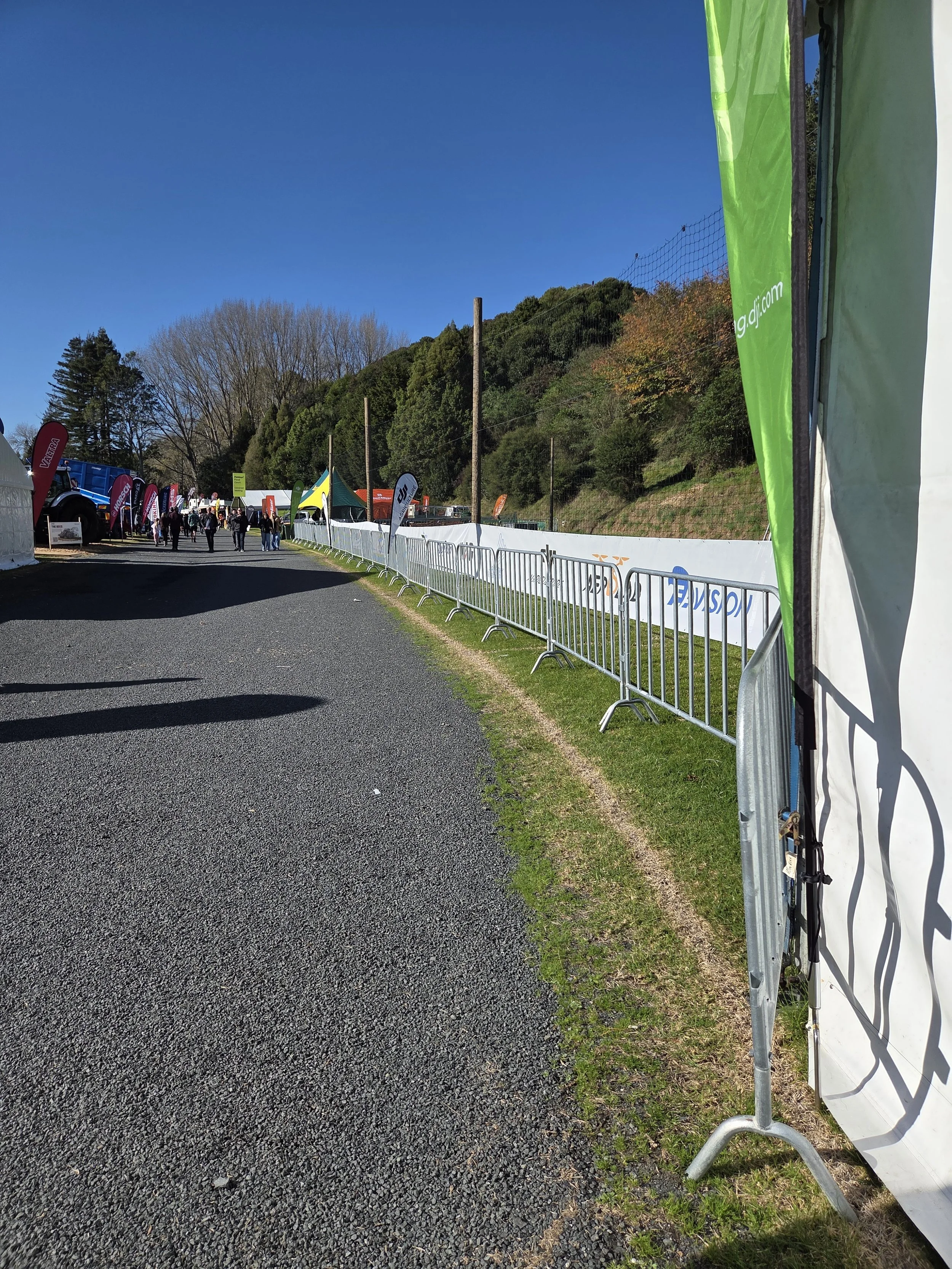 Event area with barriers, tents, and flags, set up along a road on a sunny day, with a hill covered in trees in the background.