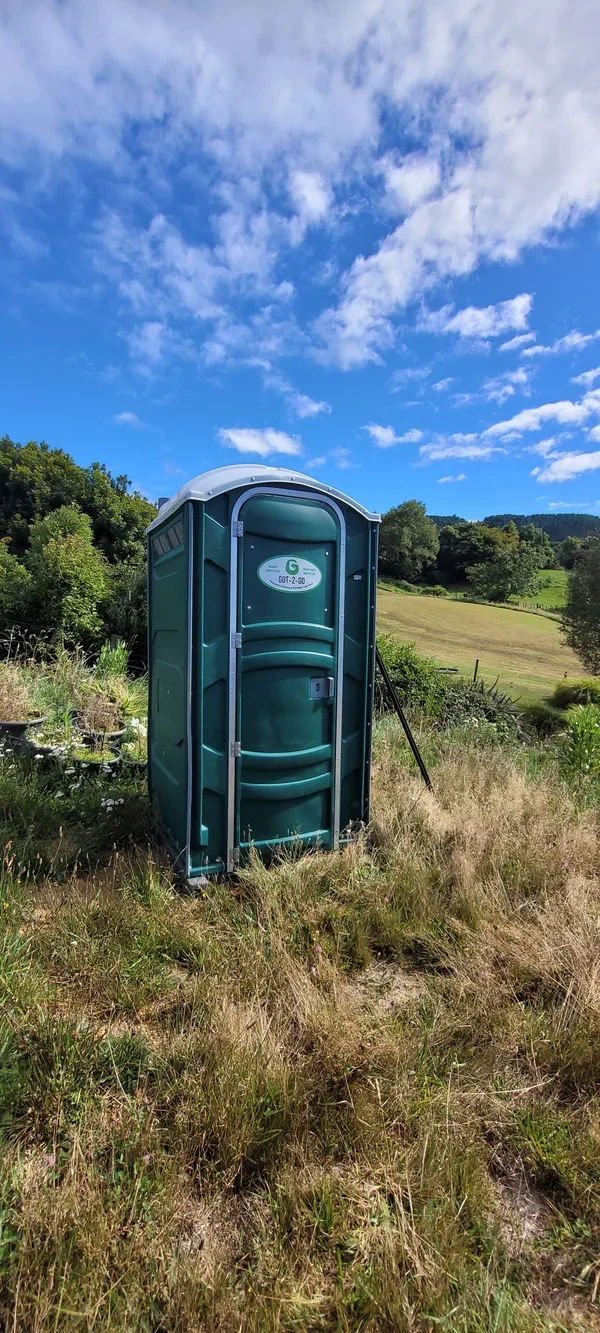 A portable toilet situated in a grassy field with trees and hills in the background, under a partly cloudy sky.