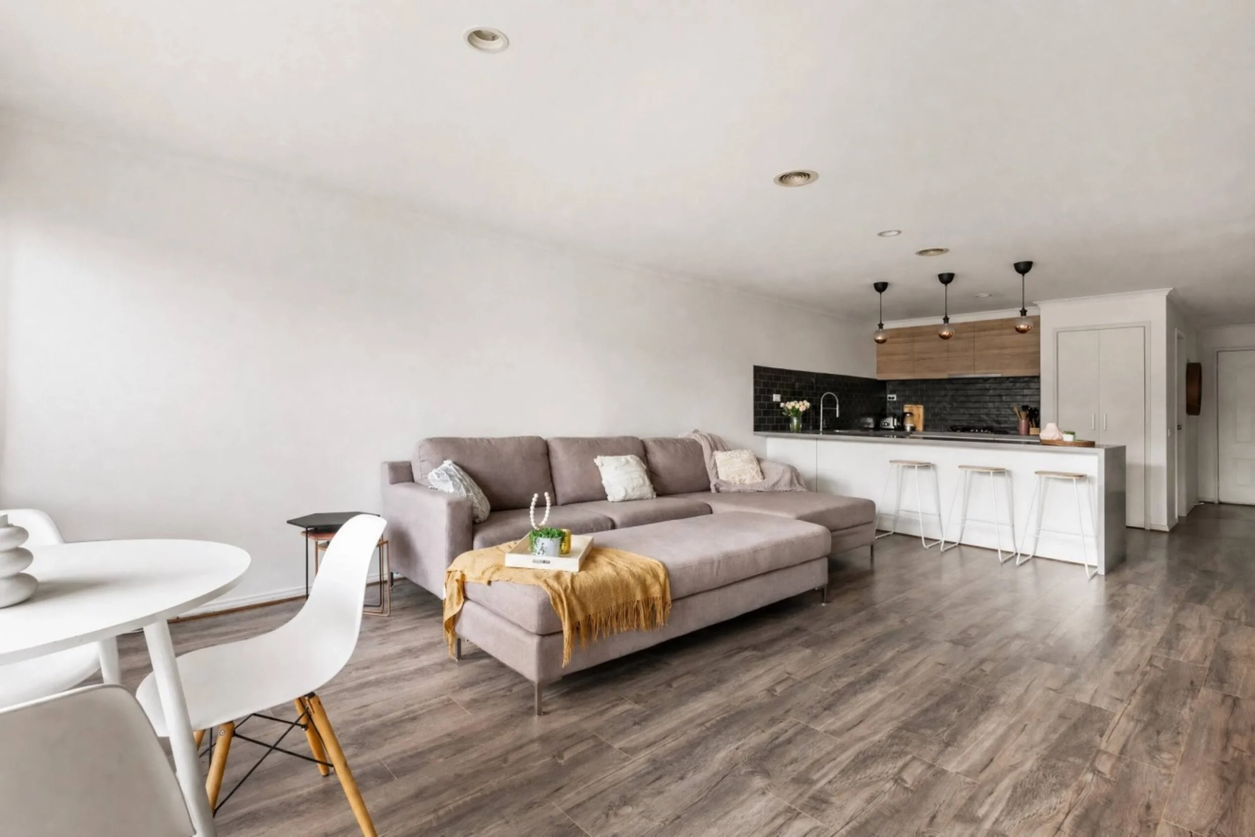 A modern living room with a gray sofa decorated with pillows, a white round dining table with white chairs, a kitchen area with black backsplash tiles, wooden cabinets, hanging pendant lights, and a breakfast bar with stools. The room has wood floori