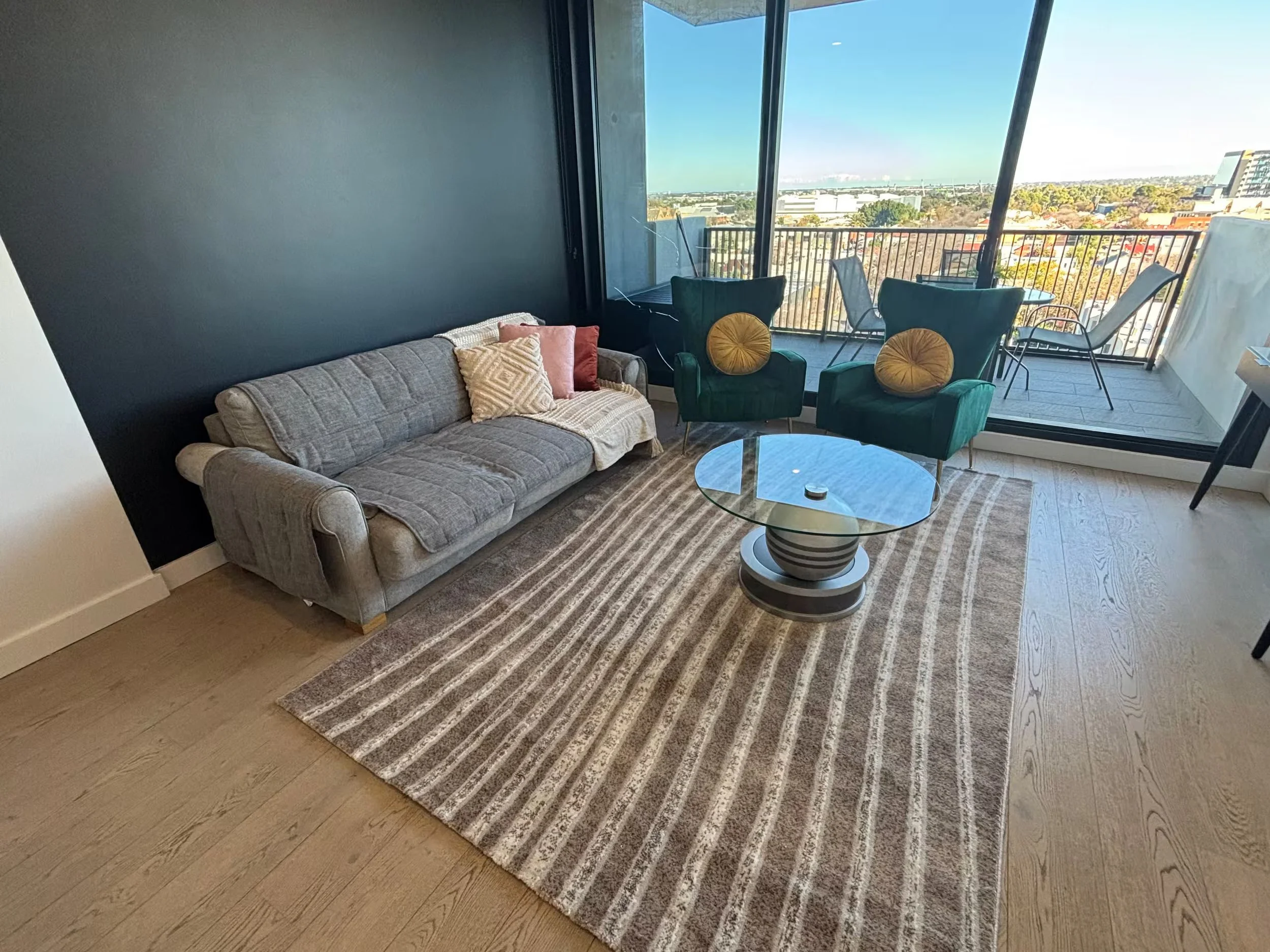 Living room with a gray couch, two green chairs, a glass coffee table, a beige and brown striped rug, and a large sliding glass door leading to a balcony with city views.