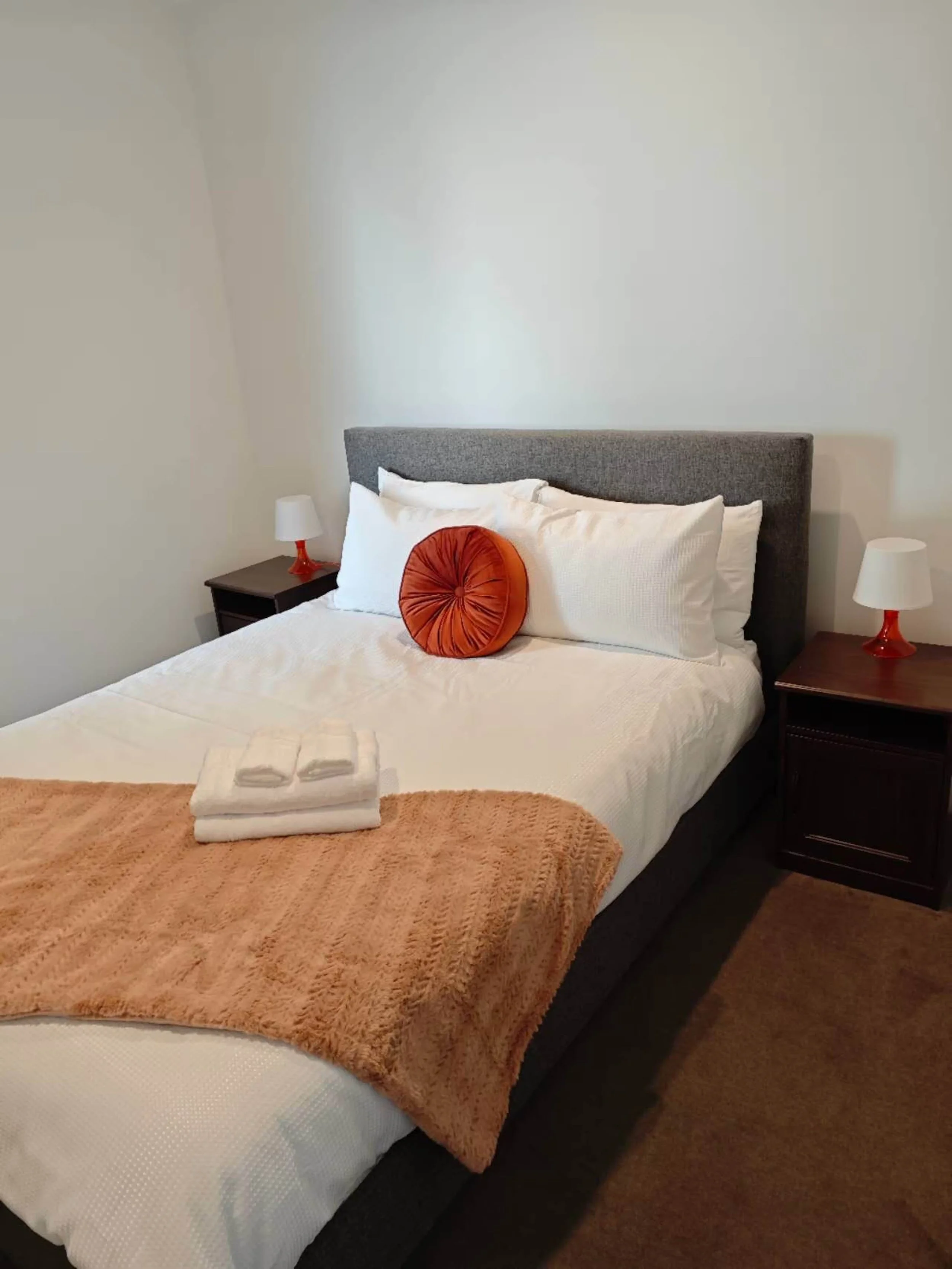 A neatly made bed with white sheets, a burnt orange decorative pillow, a folded white towel, and a brown textured blanket at the foot of the bed. Two black nightstands with white lamps and orange bases are on either side of the bed. The headboard is 