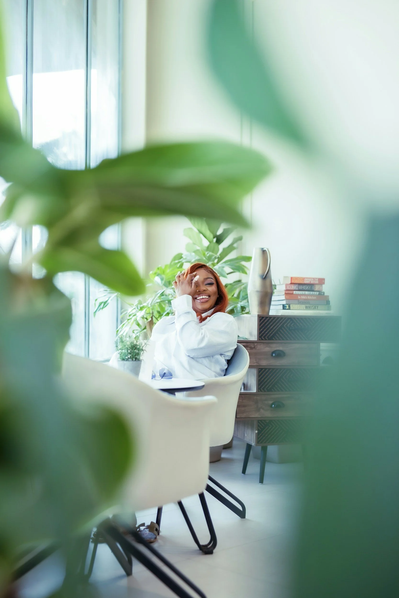 A young woman with orange hair is sitting at a desk, smiling, and touching her head. She is inside a bright, modern room with large windows and green plants. There are stacked books on a cabinet in the background.