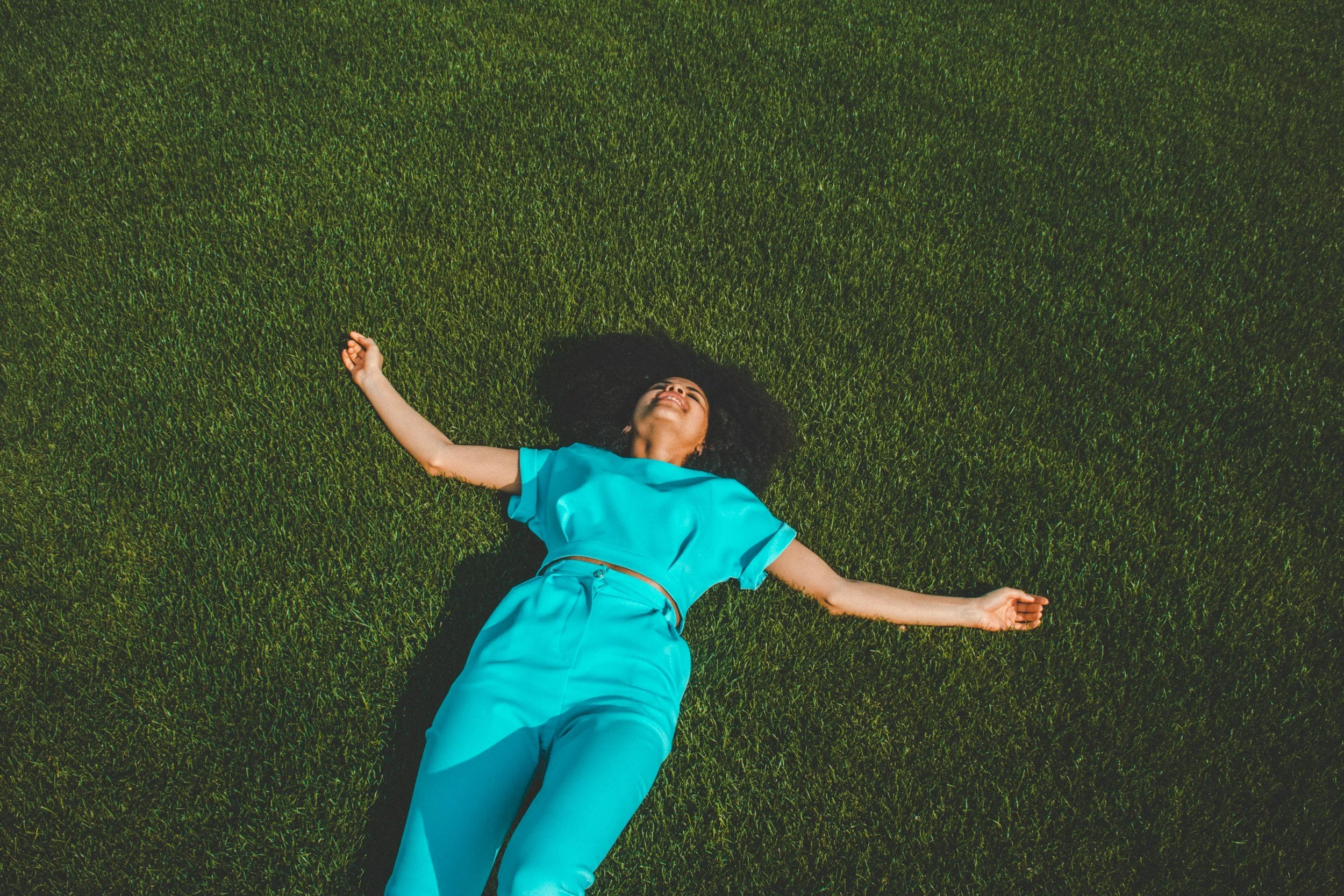 A woman with curly black hair lying on her back on a lush green grassy field, smiling and wearing a bright blue shirt and matching pants.