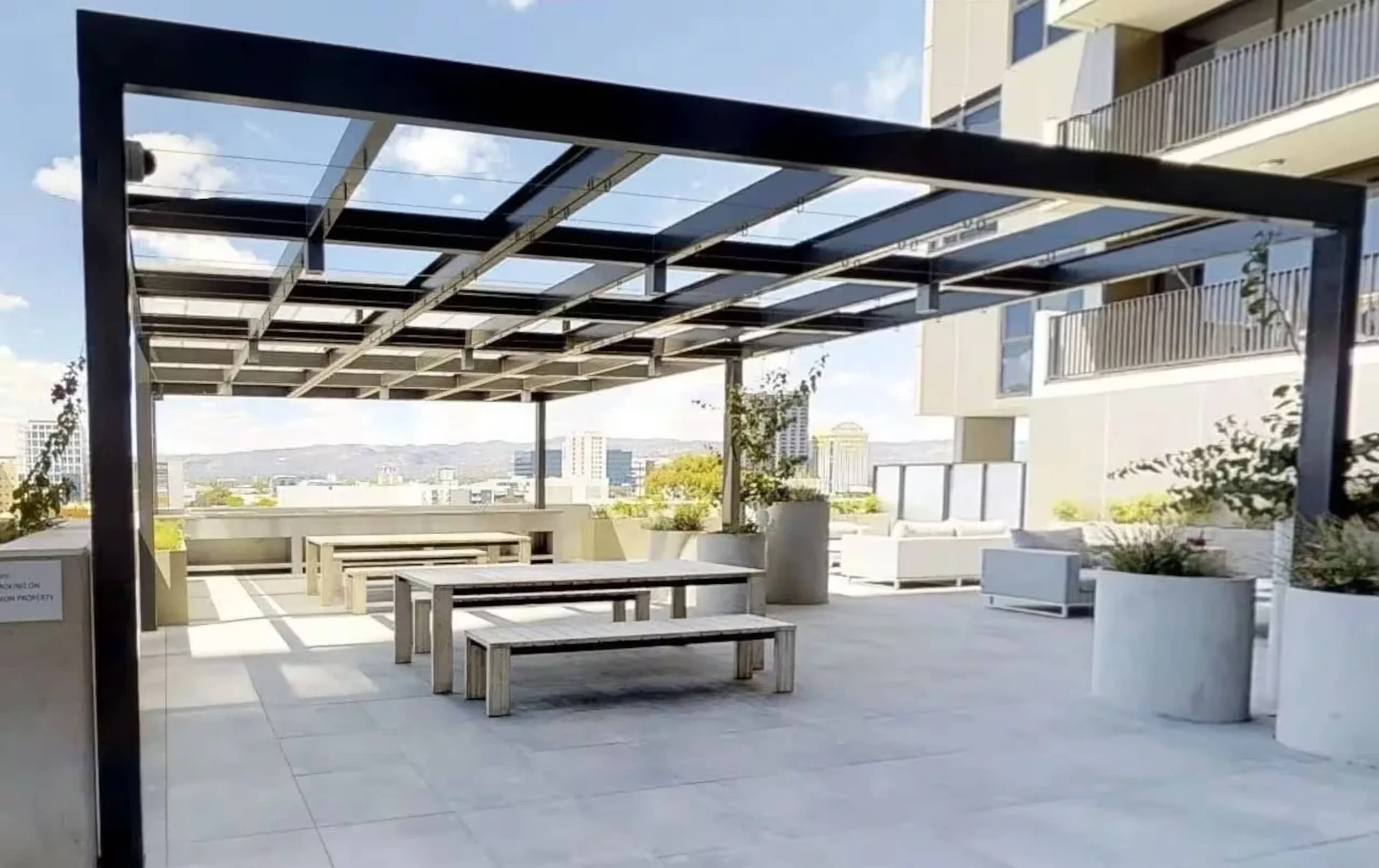 A rooftop terrace with a modern pergola structure made of black metal and glass panels, overlooking a cityscape with buildings and mountains in the distance.