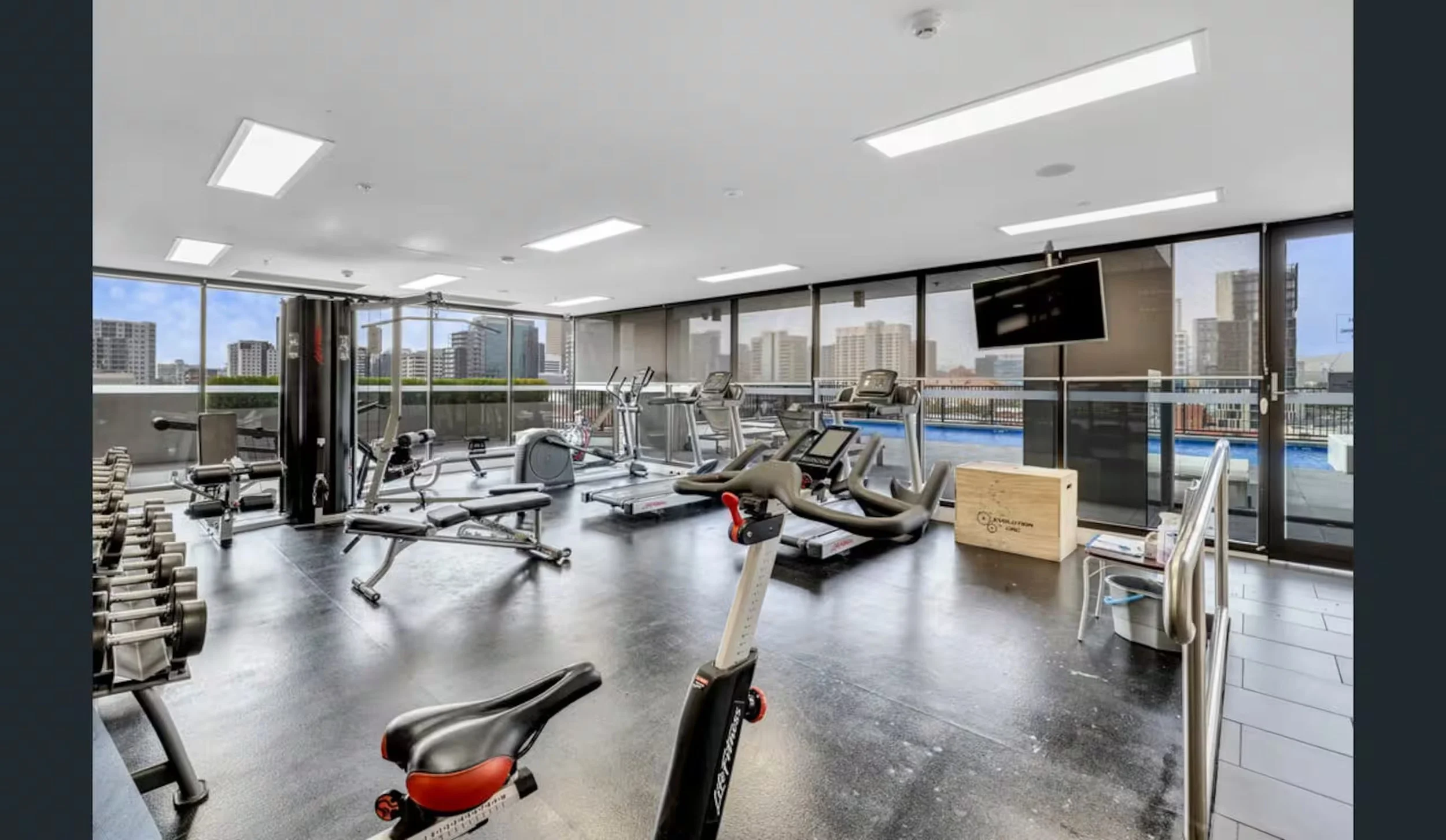 A modern gym with various exercise equipment, including treadmills, stationary bikes, and free weights, set against large windows with a city skyline view.
