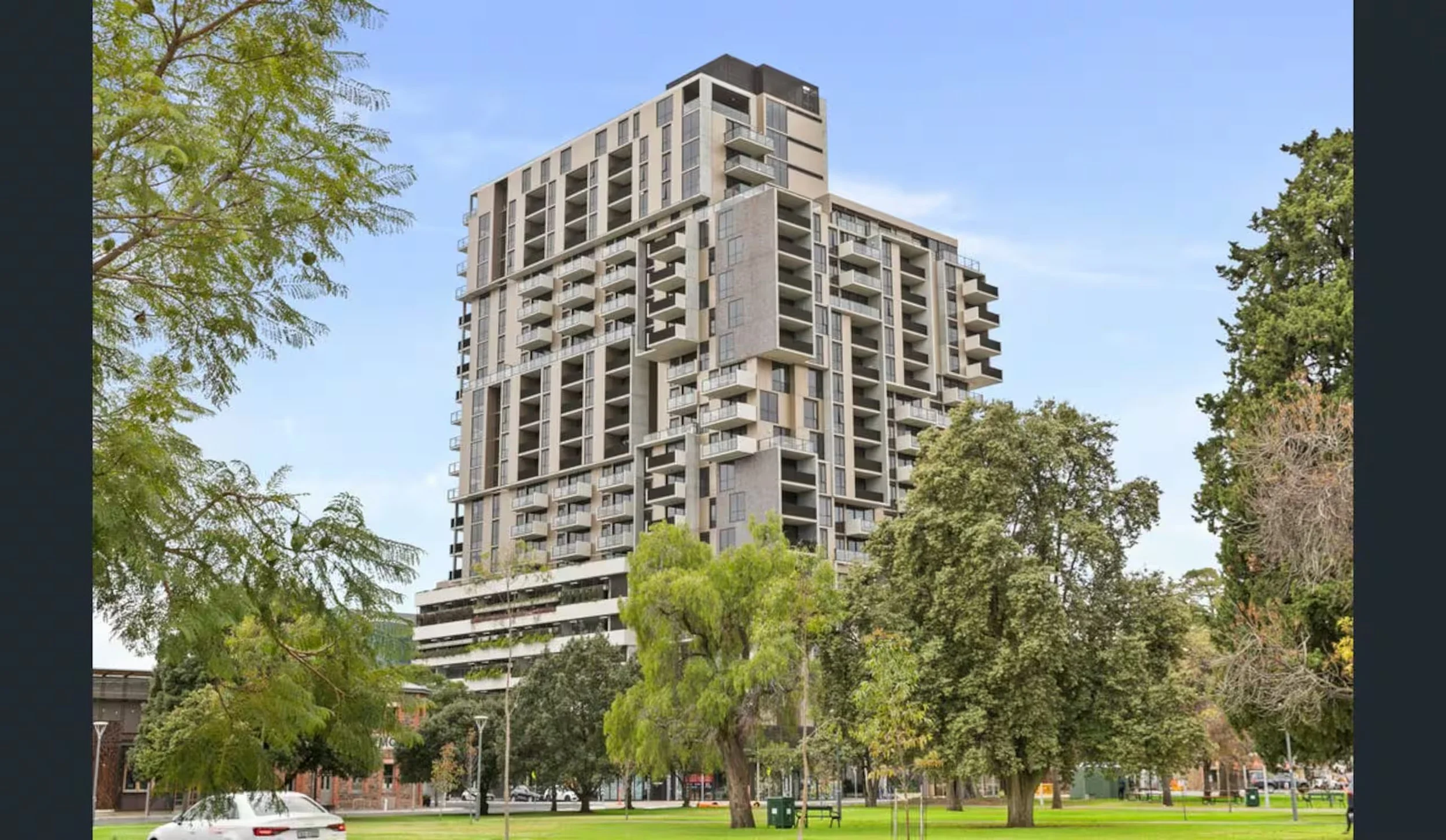 A modern high-rise apartment building surrounded by trees in a park with benches and a street with cars in the foreground.