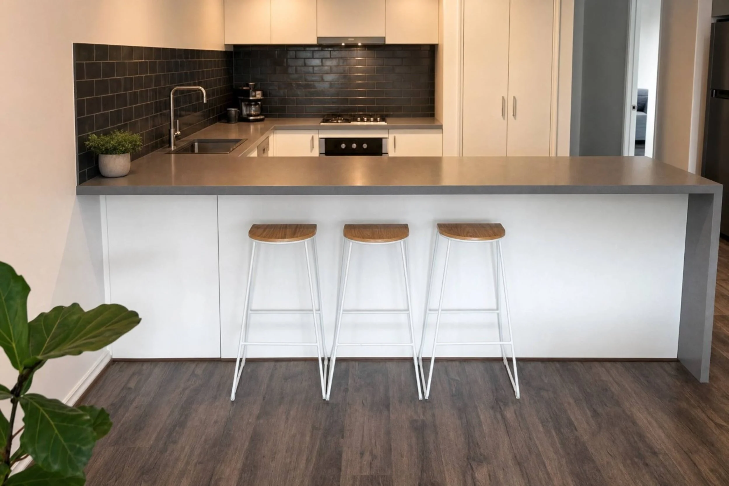 A modern kitchen with a beige countertop island, three wooden stools with white legs, black subway tile backsplash, white cabinets, a coffee maker, and a stainless steel oven. Part of a living room is visible through a doorway on the right.