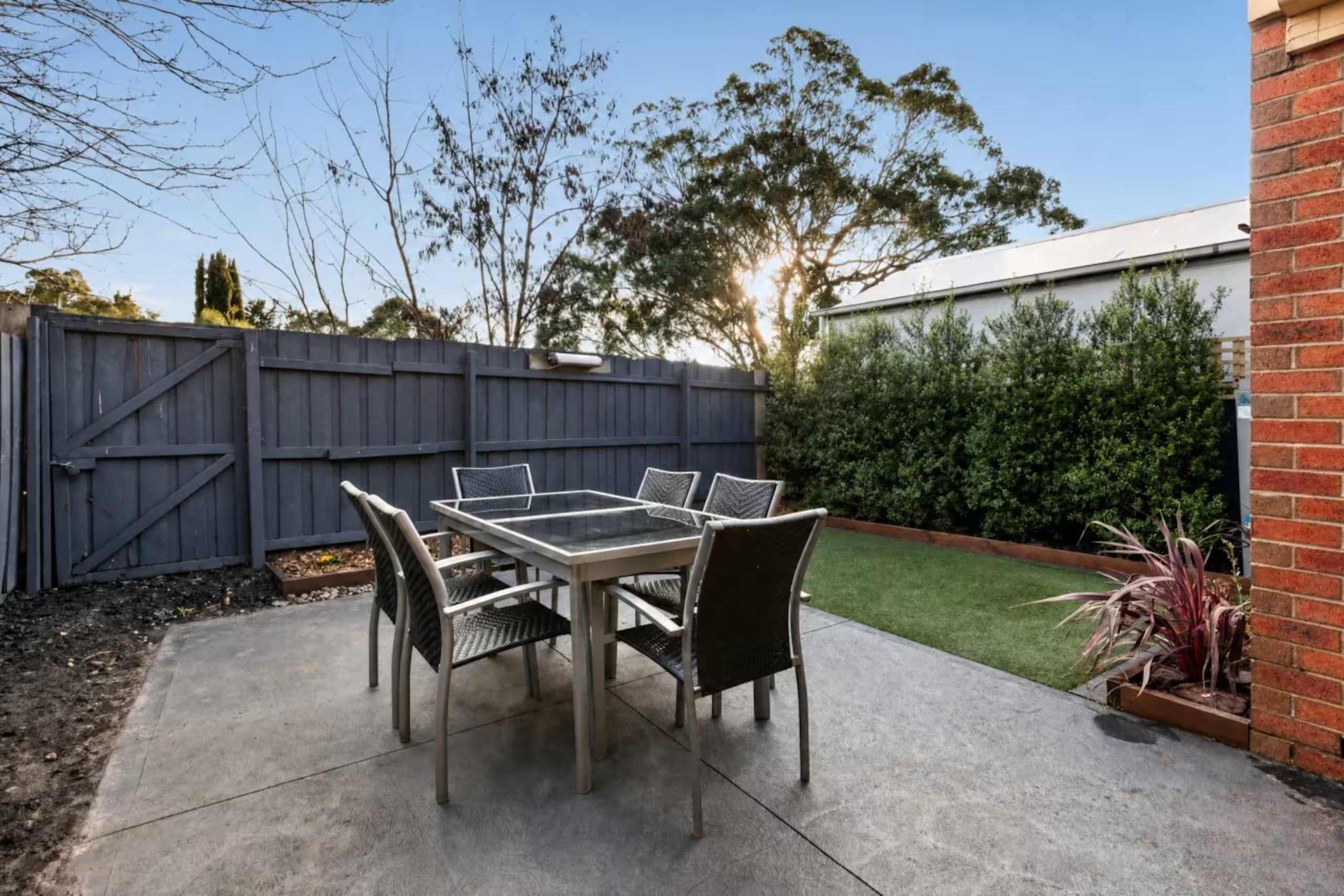 An outdoor patio area with a glass table and six chairs on a concrete slab. There is a gray wooden fence in the background, with trees and bushes behind it. The sun is shining through the trees, indicating that it is either early morning or late afte