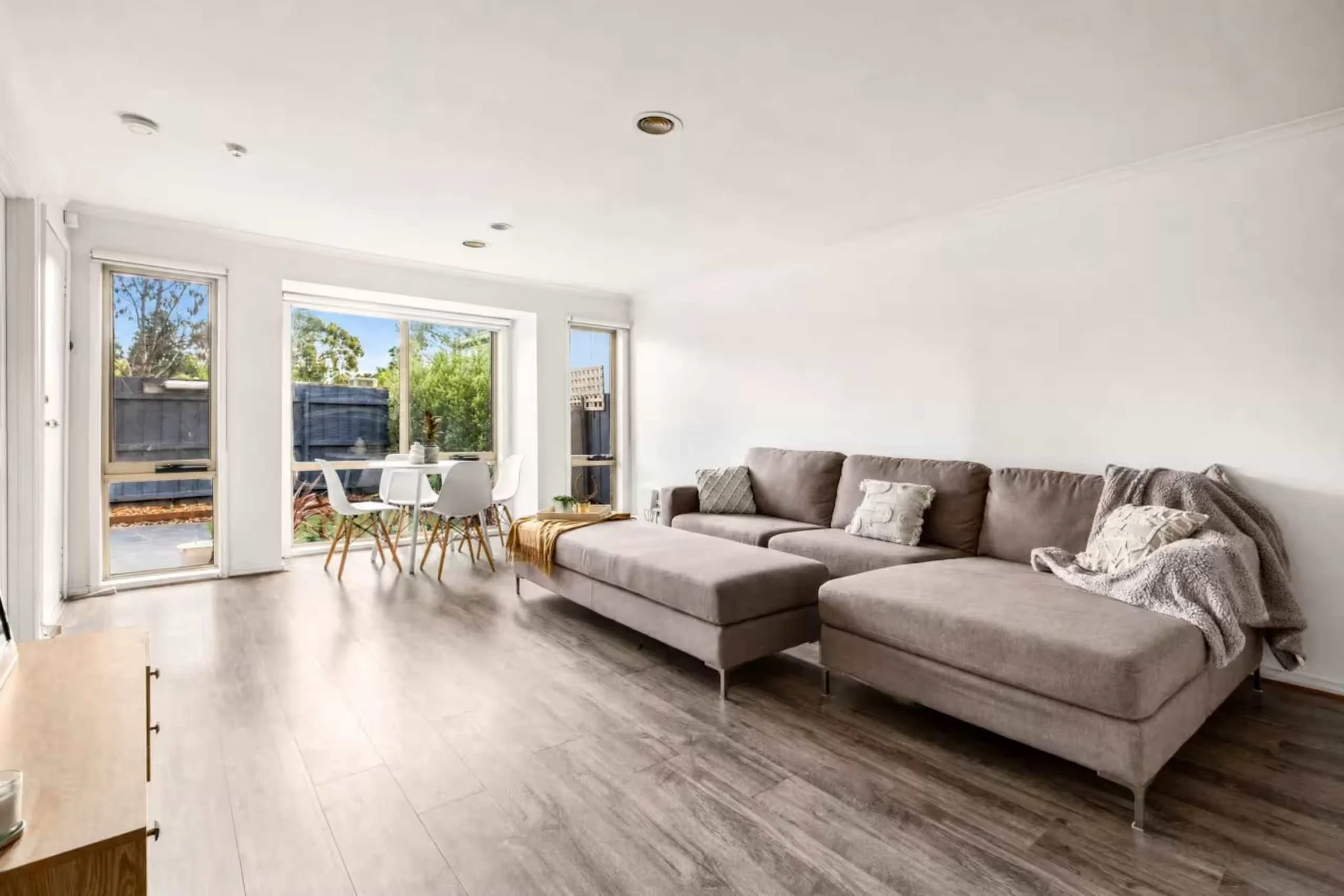 Living room with gray sectional sofa, decorative pillows, throw blanket, light-colored wood flooring, large windows with views of trees and backyard, and some small furniture pieces.