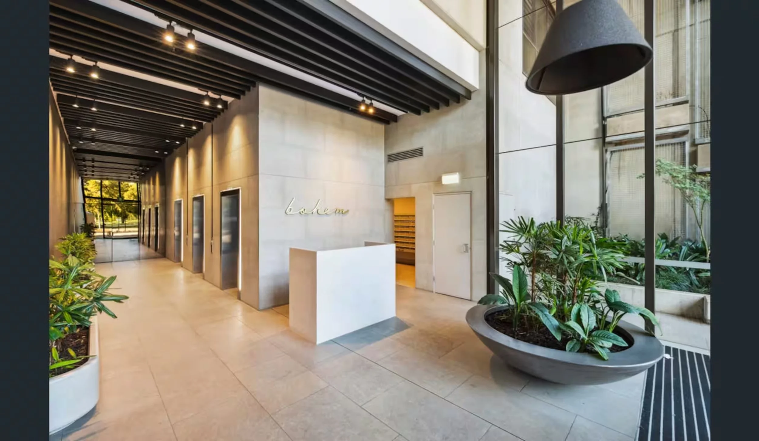 Modern lobby with beige tile floor, large potted plants, and floor-to-ceiling windows with greenery outside, black ceiling beams, and a white reception desk.