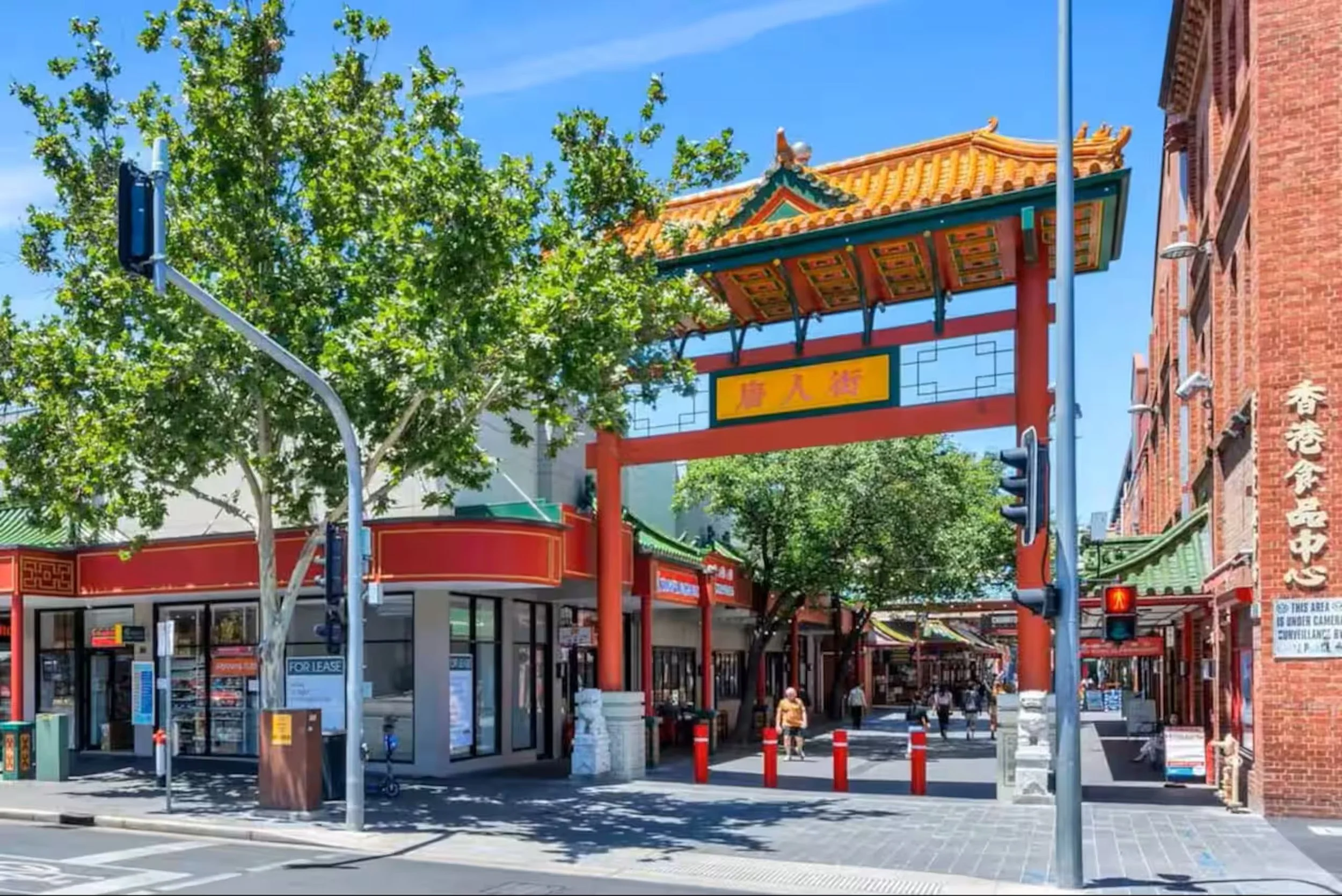 A Chinatown entrance gate with Chinese architectural features, red columns, and a decorative roof, in an urban setting with trees and buildings nearby, under a blue sky.