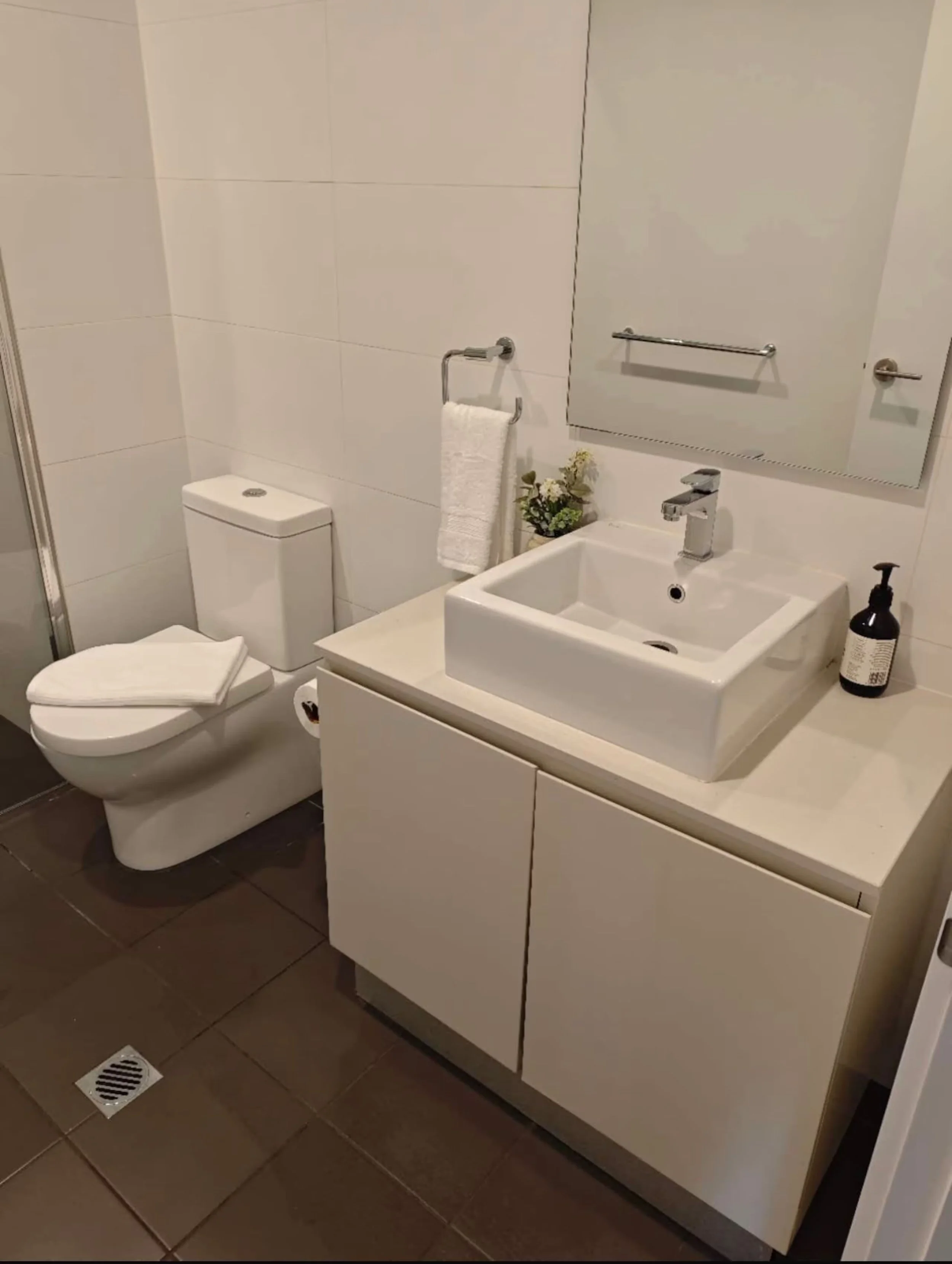 A modern bathroom with white tiled walls, a white toilet, a white vanity with a sink, a large mirror, and a small potted plant on the counter. There is a towel hanging on a wall-mounted towel bar and a black soap dispenser on the sink. The floor is t