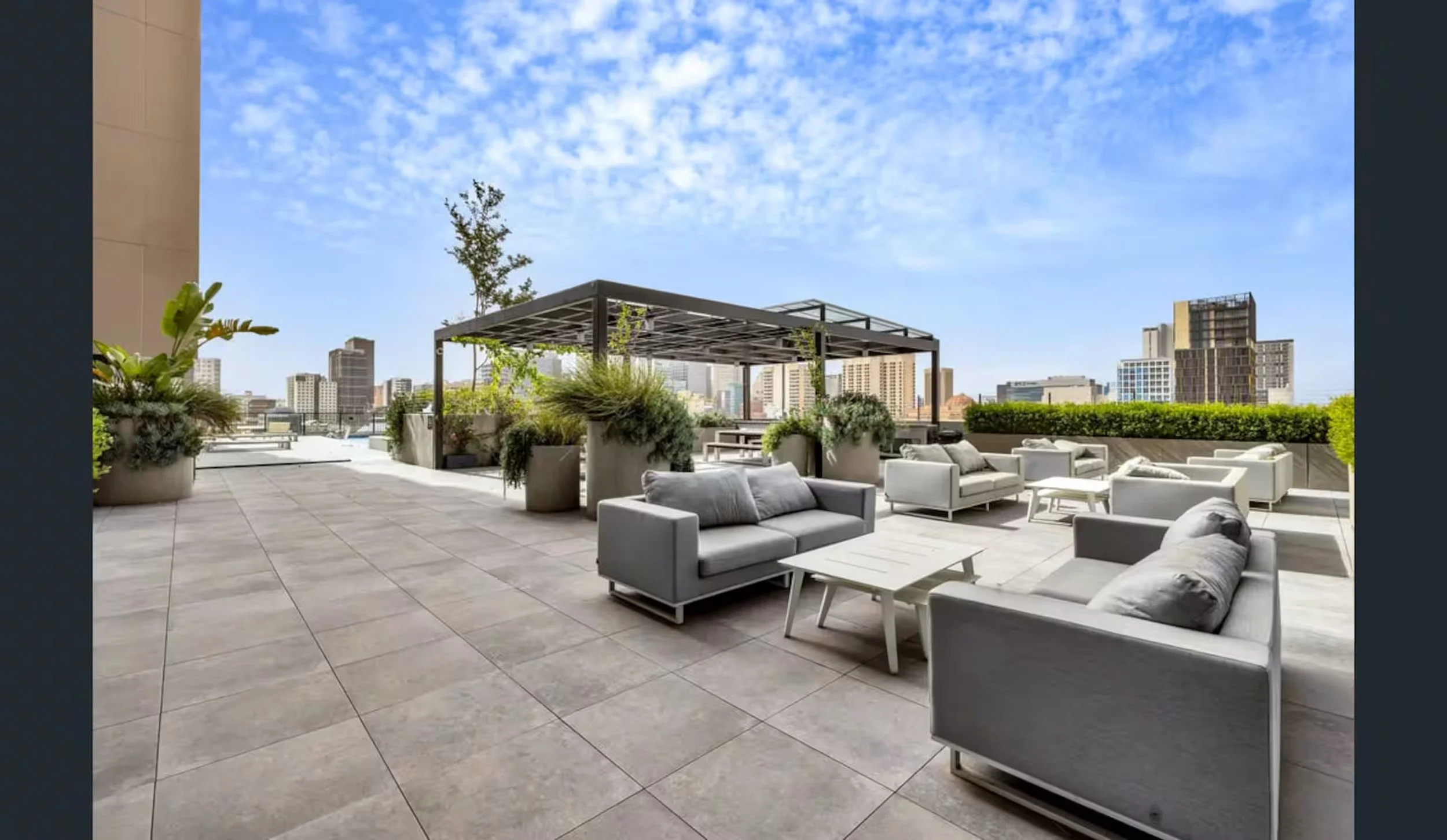 Rooftop patio with outdoor furniture including sofas and chairs, potted plants, a pergola, and a city skyline in the background under a partly cloudy sky.