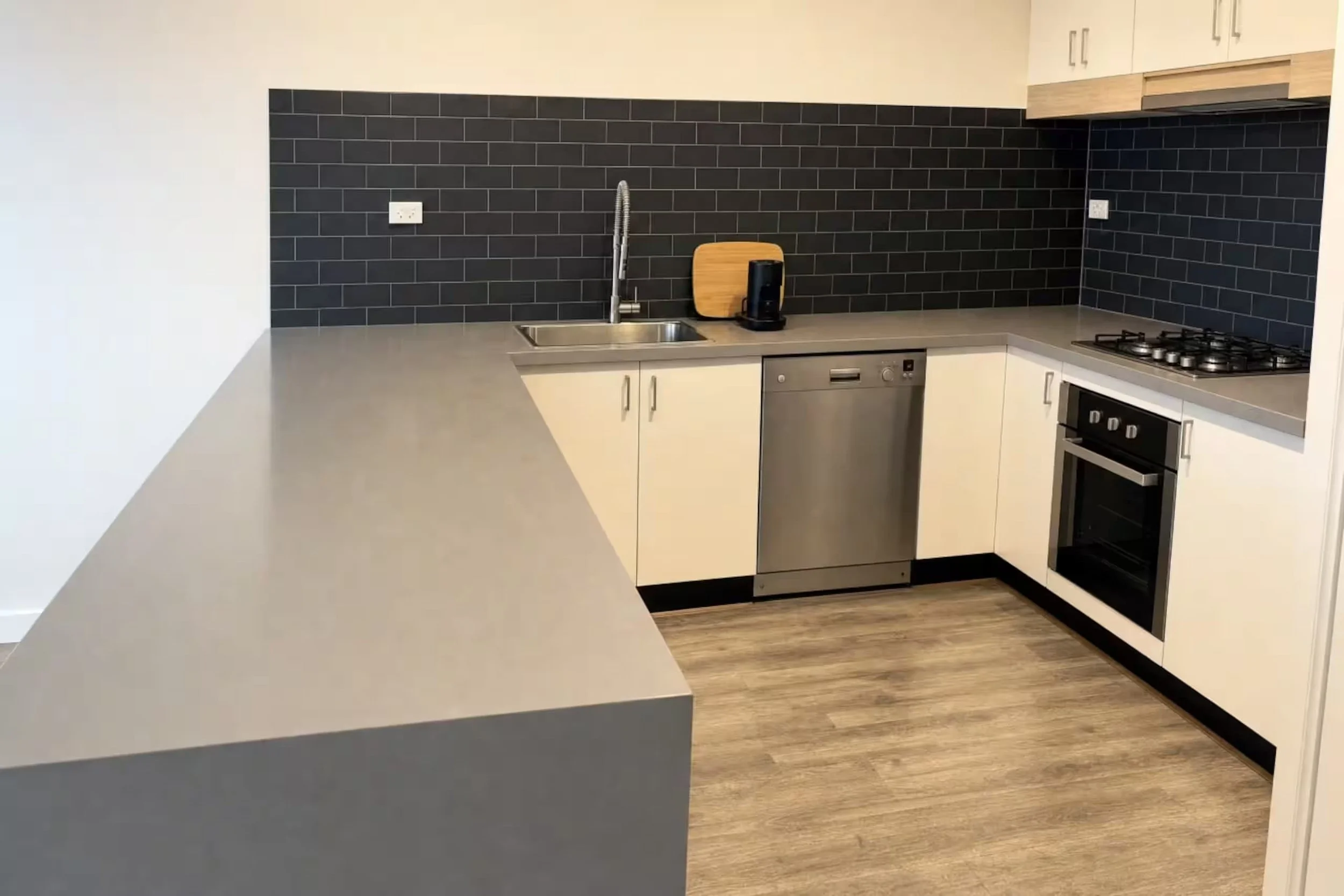 Modern kitchen with black subway tile backsplash, white cabinets, and stainless steel appliances including a dishwasher and oven. There is a gray countertop with a sink and a coffee machine on it.