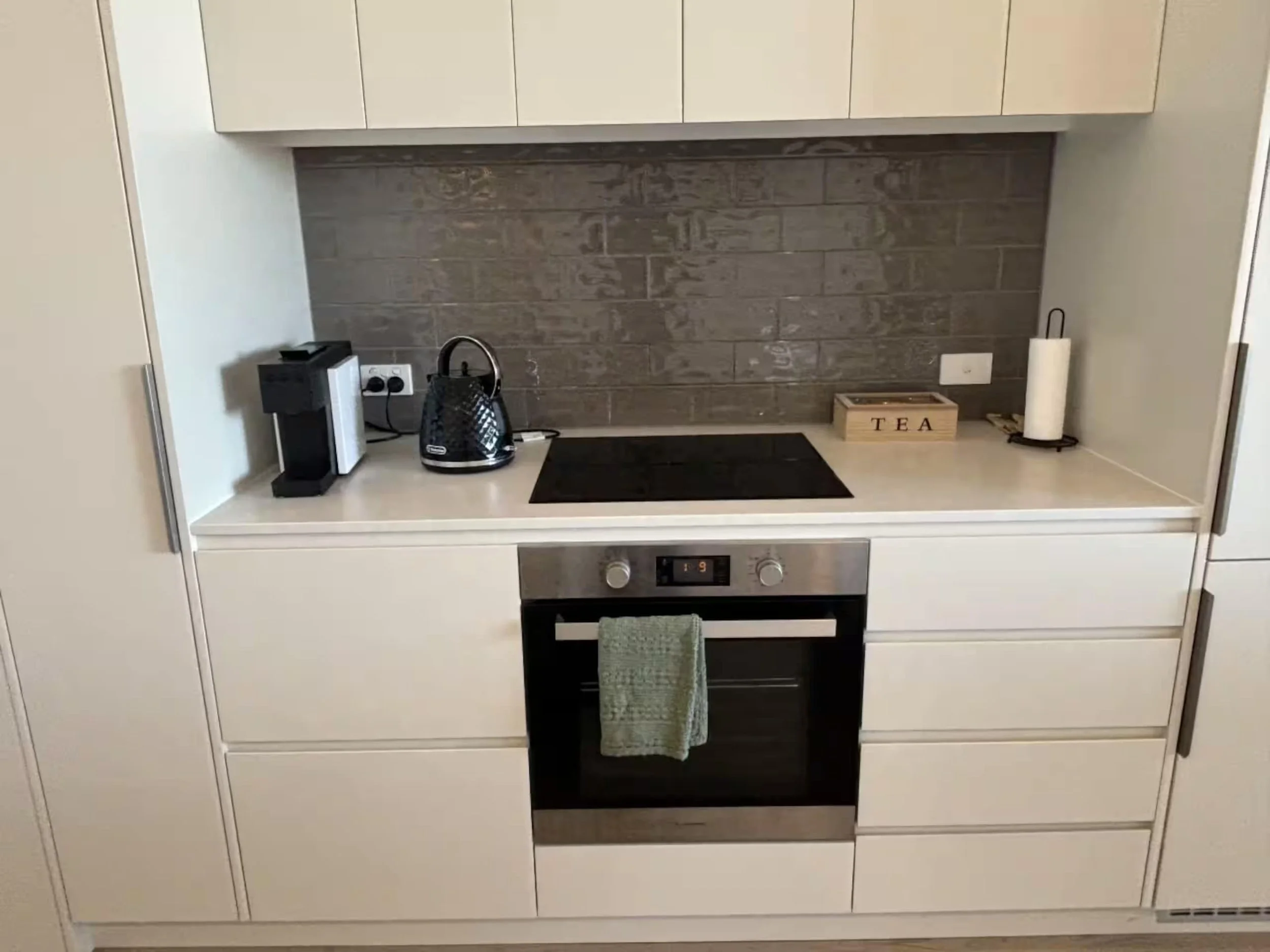 A modern kitchen with white cabinets and a beige countertop. There is a black and silver electric kettle, a small black and white appliance, a wooden box labeled 'TEA', a paper towel roll, and a black oven with a green towel hanging from its handle. 