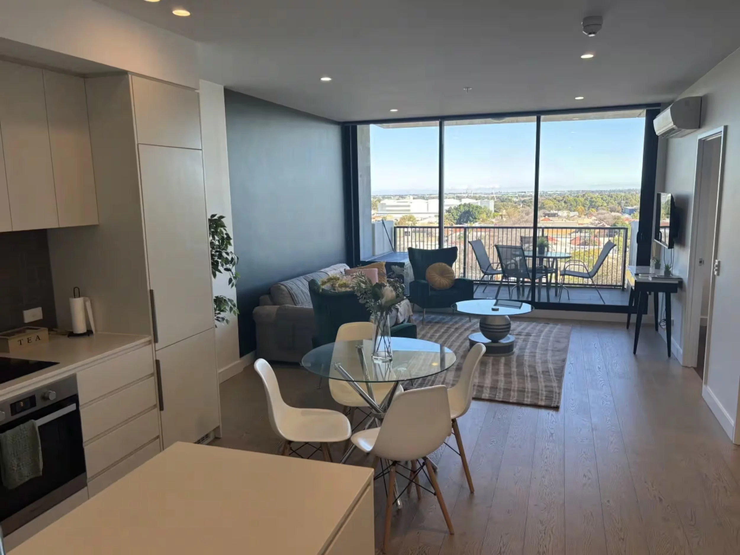 Modern apartment interior with open living space, large floor-to-ceiling windows overlooking a cityscape, a cozy gray sofa with decorative pillows, a round coffee table, a dining area with a round glass table and white chairs, and a balcony with outd