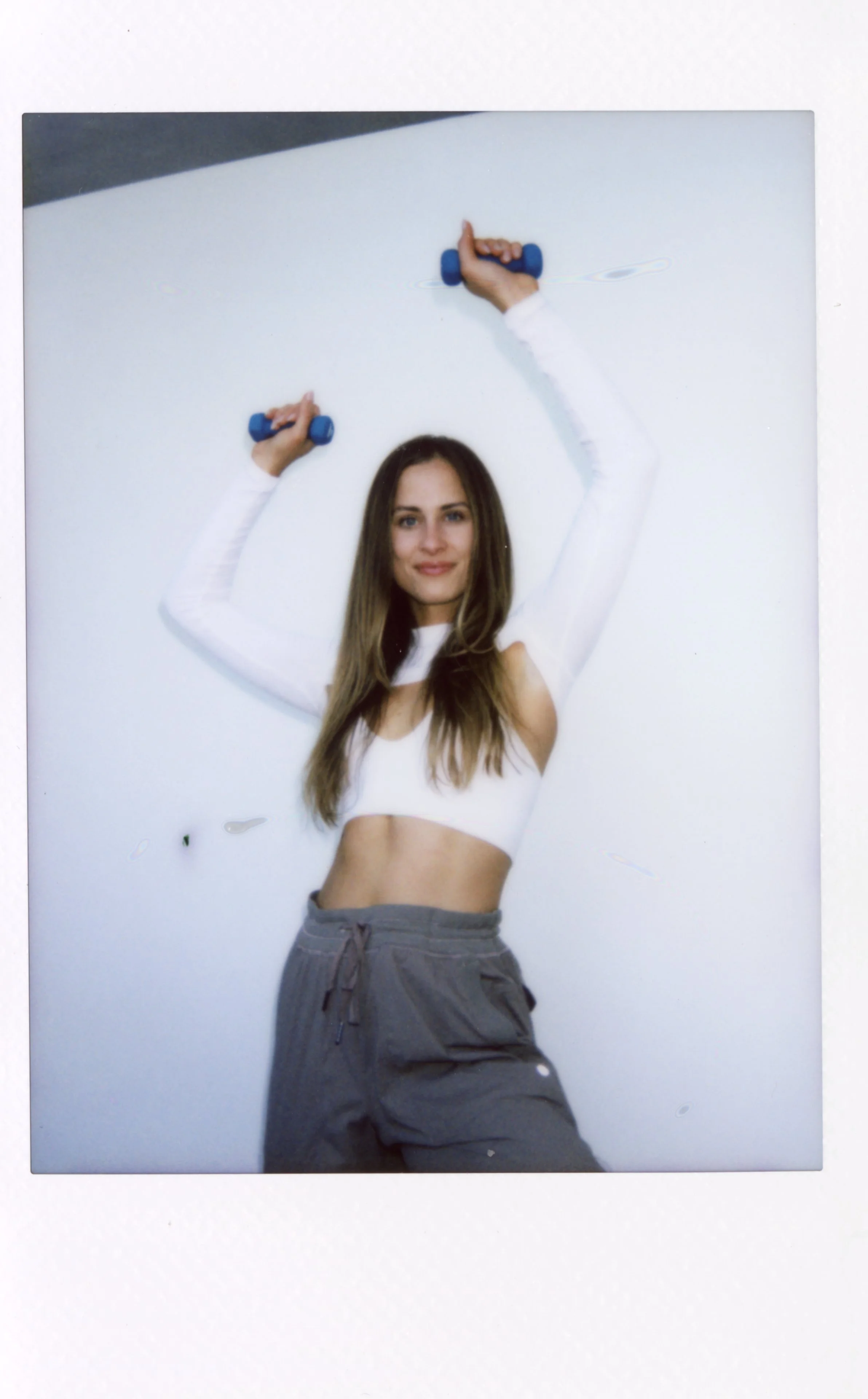 Young woman in white long-sleeve crop top and gray sweatpants holding small blue dumbbells above her head against a white wall.