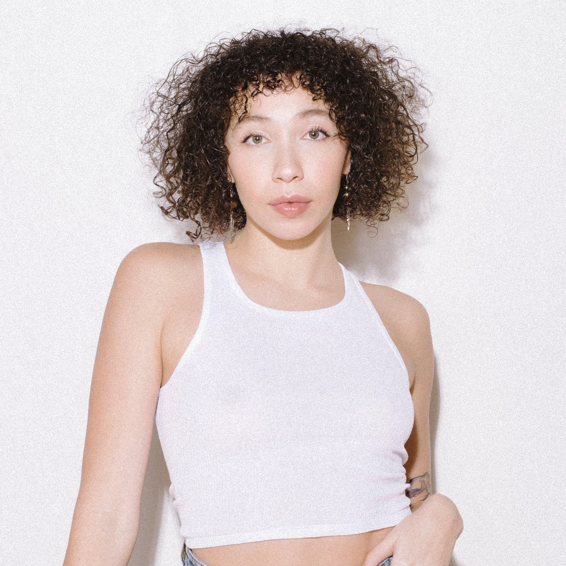 A young woman with curly brown hair wearing a white sleeveless top and hoop earrings standing against a plain white background.