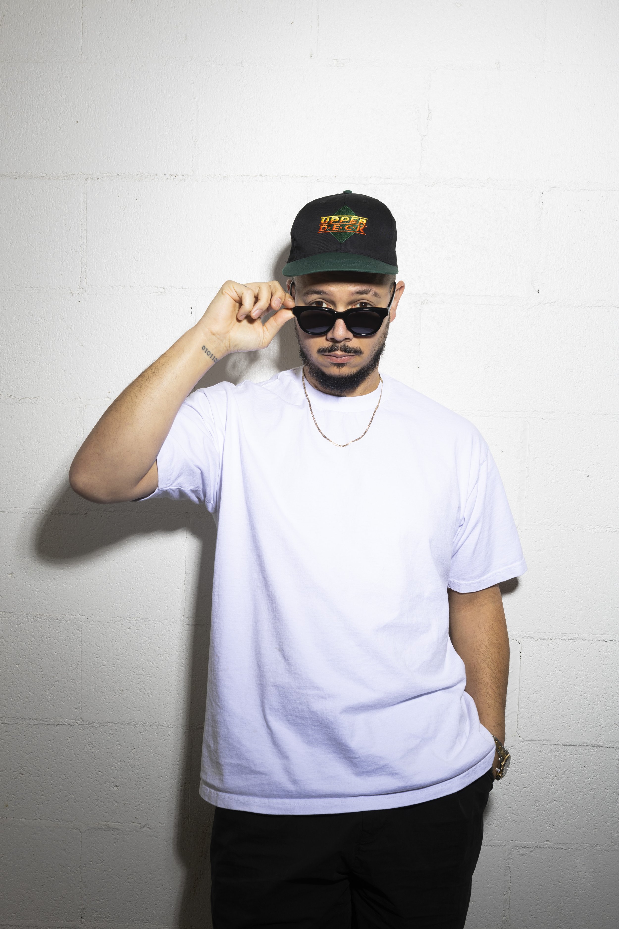 A man wearing sunglasses, a black cap with colorful text, a white t-shirt, a gold chain necklace, black pants, and a watch standing against a white wall.