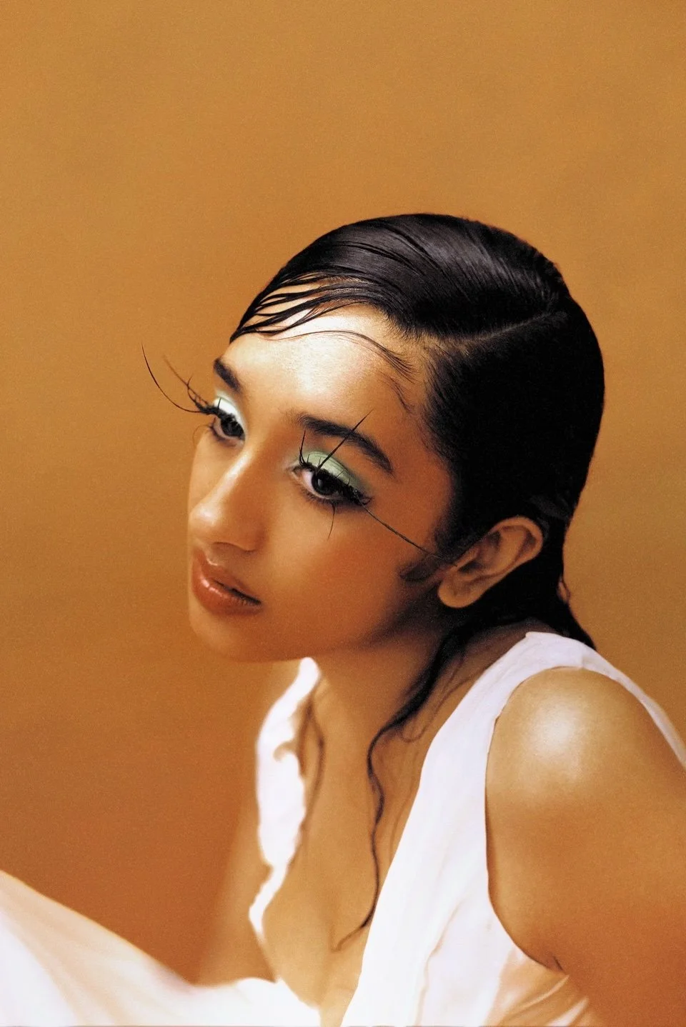 Close-up portrait of a woman with slicked-back black hair, green eyeshadow, and long, stylized eyelashes, wearing a white top against a warm beige background.