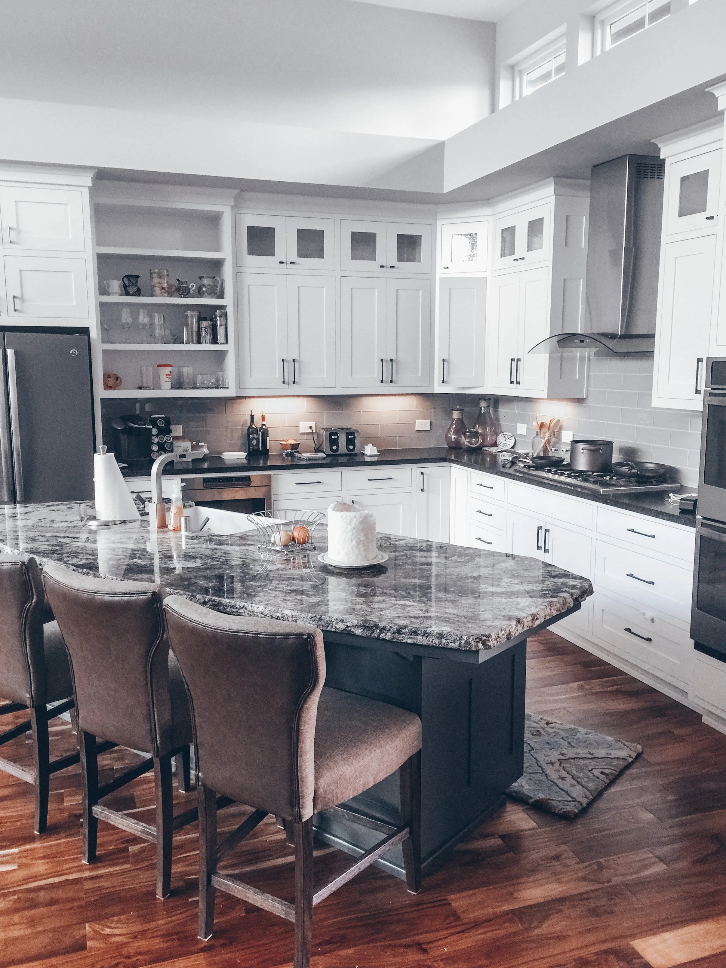 A modern kitchen with white cabinets, a granite island with four brown chairs, stainless steel appliances, and a hardwood floor.