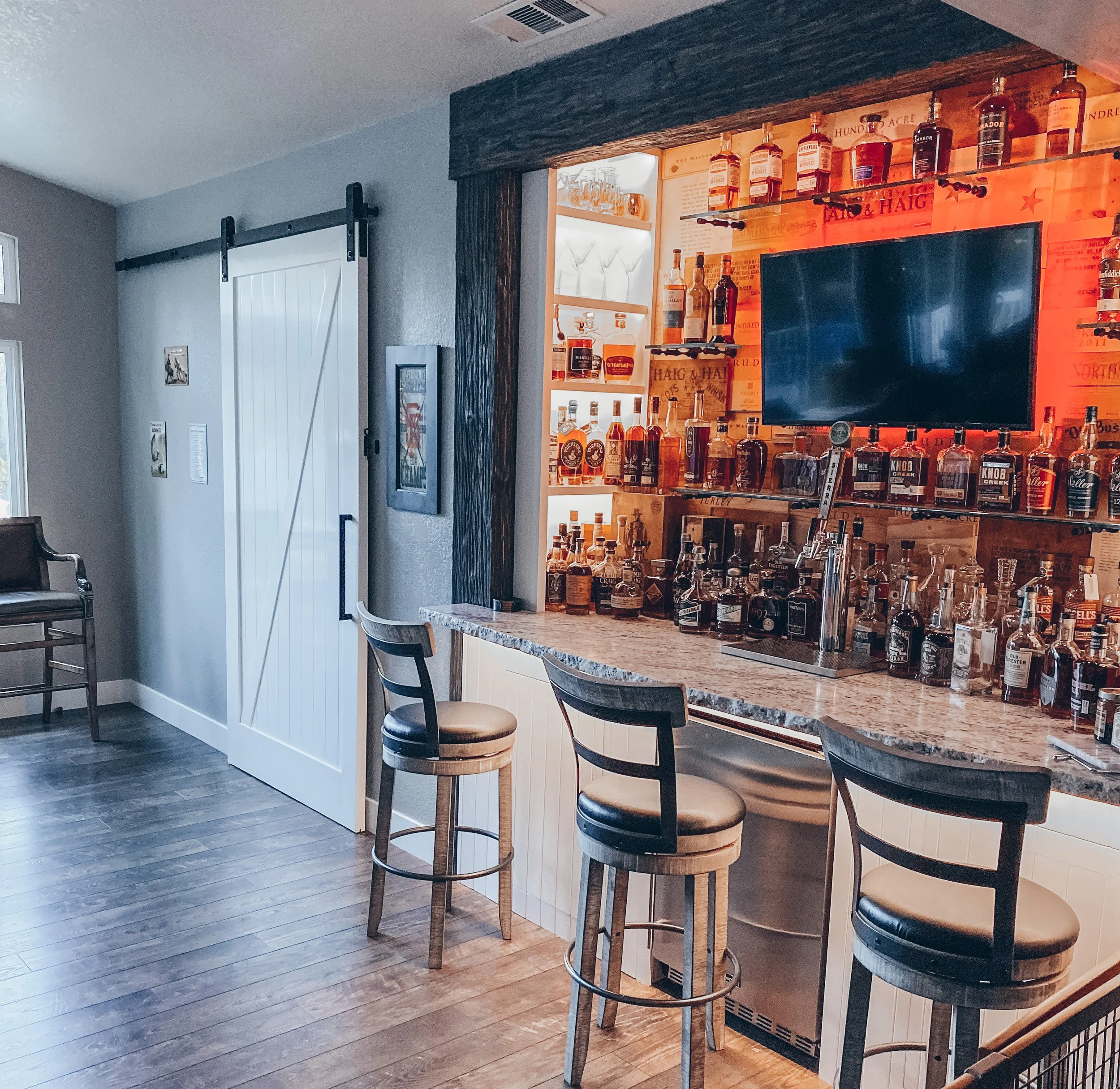 A home bar with a granite countertop, wooden bar stools, and shelves stocked with various bottles of alcohol. A television is mounted on the alcohol shelf area, with warm lighting behind the bottles.
