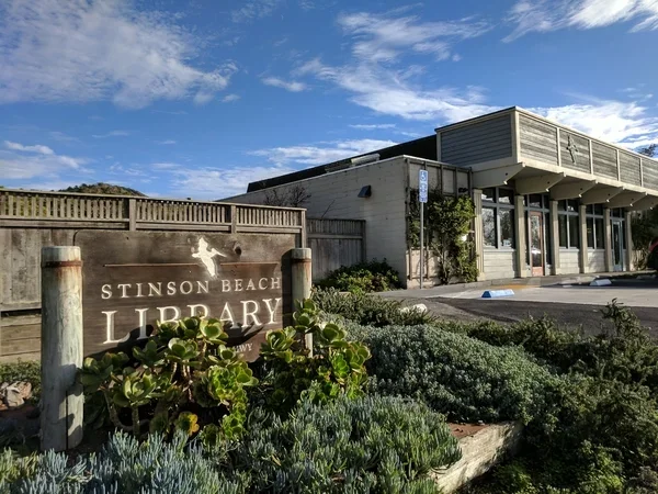 Exterior view of the Stinson Beach Library building with a landscaped area in front, including plants and a wooden sign that reads 'Stinson Beach Library' with a silhouette of a bird.
