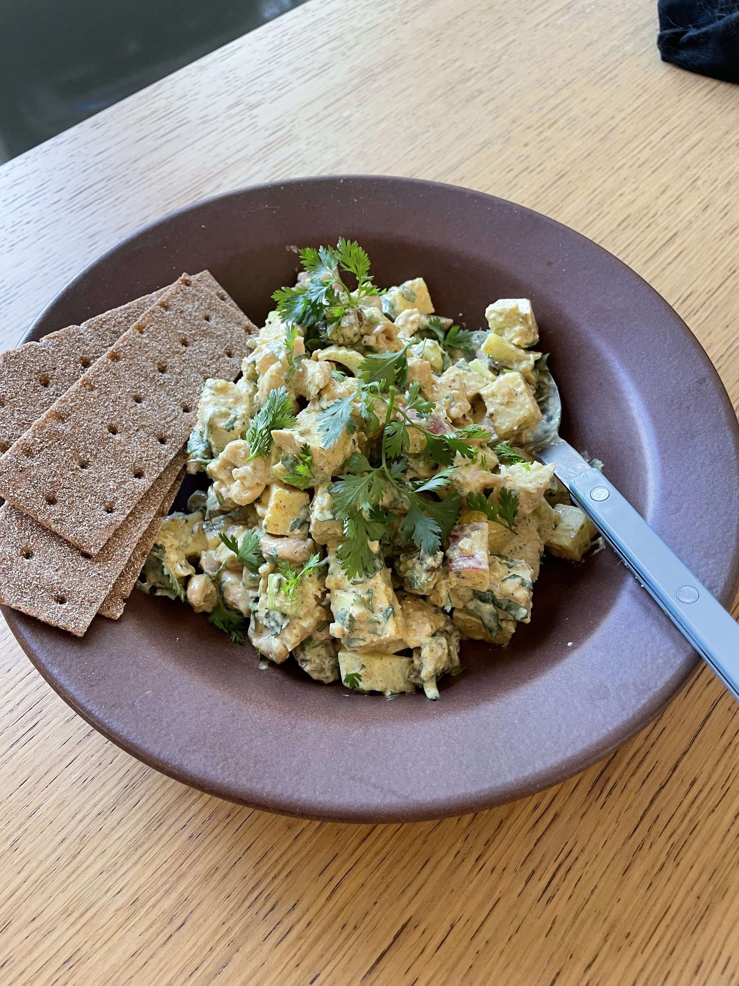 A brown bowl containing potato salad garnished with cilantro and served with whole grain crackers on a wooden table.