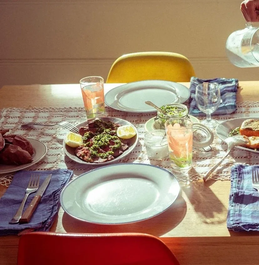 A table set for a meal with plates, utensils, glasses, a bowl of meat with lemon wedges, and a jar of green sauce, illuminated by sunlight.