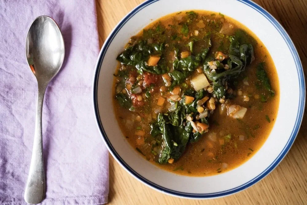 Bowl of vegetable soup with greens, chopped vegetables, and broth, on a wooden table with a soap and purple cloth nearby. postpasrtum. healthy. breastfeeding. nutrient dense.