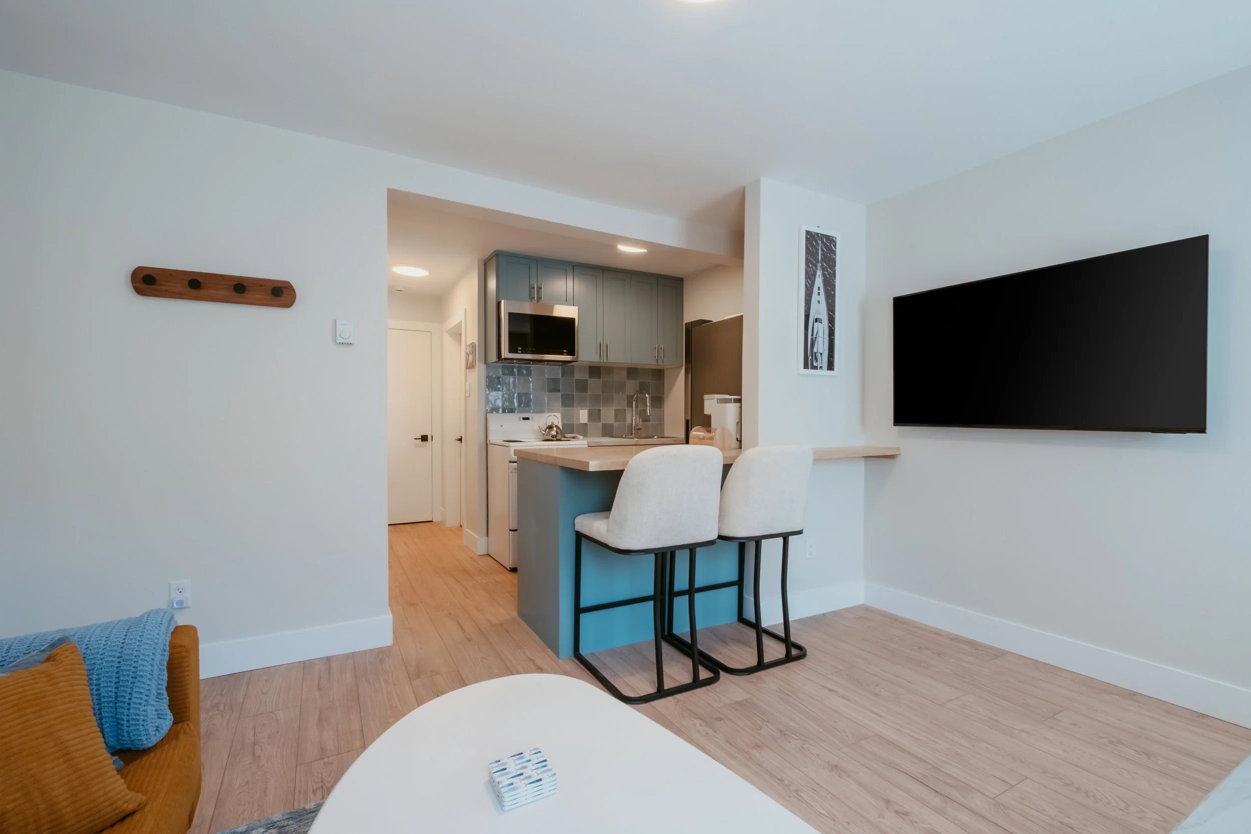 Living room with a wall-mounted flat-screen TV, a small kitchen area with gray cabinets, a microwave, a stove, a tiled backsplash, two white bar stools at a breakfast bar, and a colorful sofa with throw blankets.