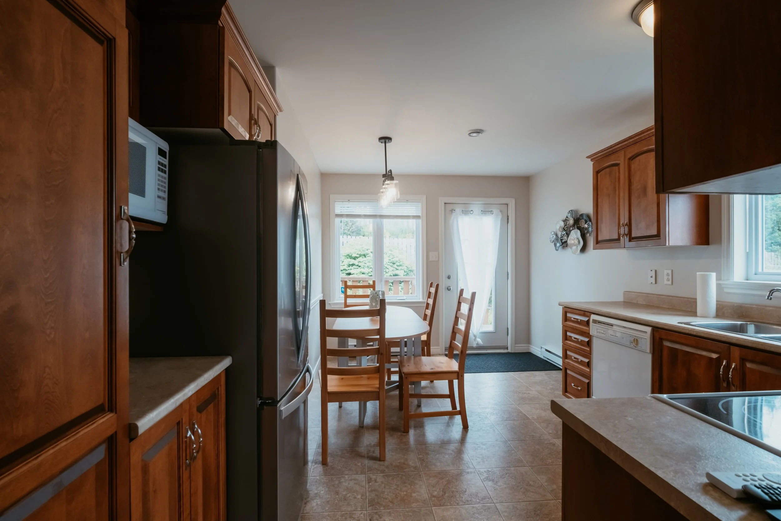 View of a kitchen with wooden cabinets, a microwave, a refrigerator, a dining table with chairs, and a door with a window leading outside. Natural light filters in through the windows.