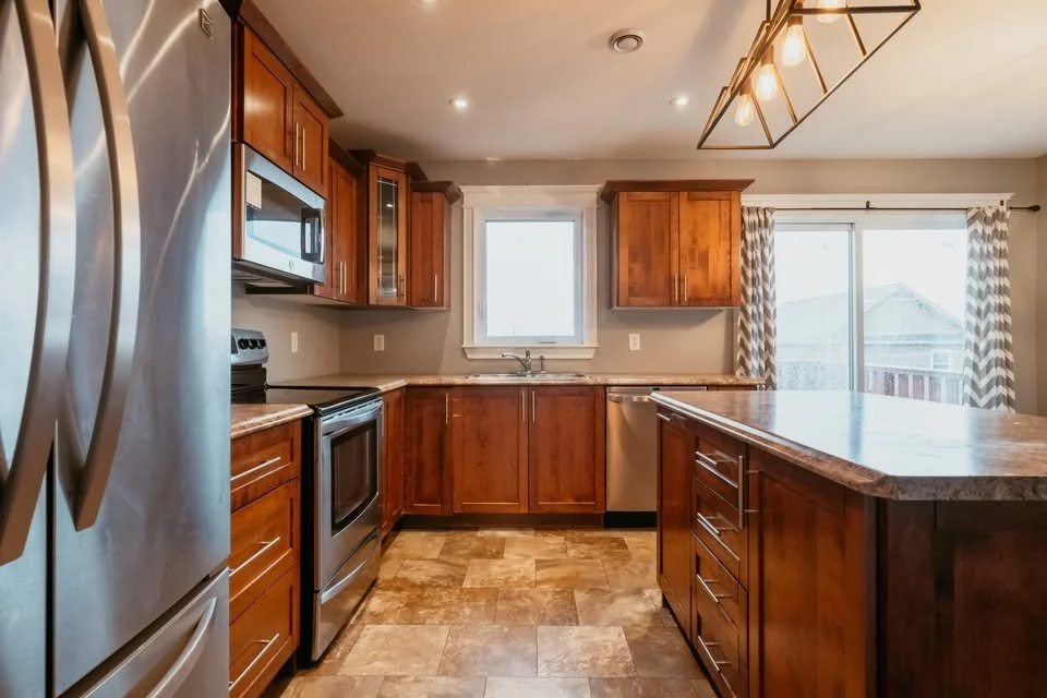 Modern kitchen with wooden cabinets, stainless steel appliances, and a central island. There are windows with curtains and a dining area with a hanging light fixture.