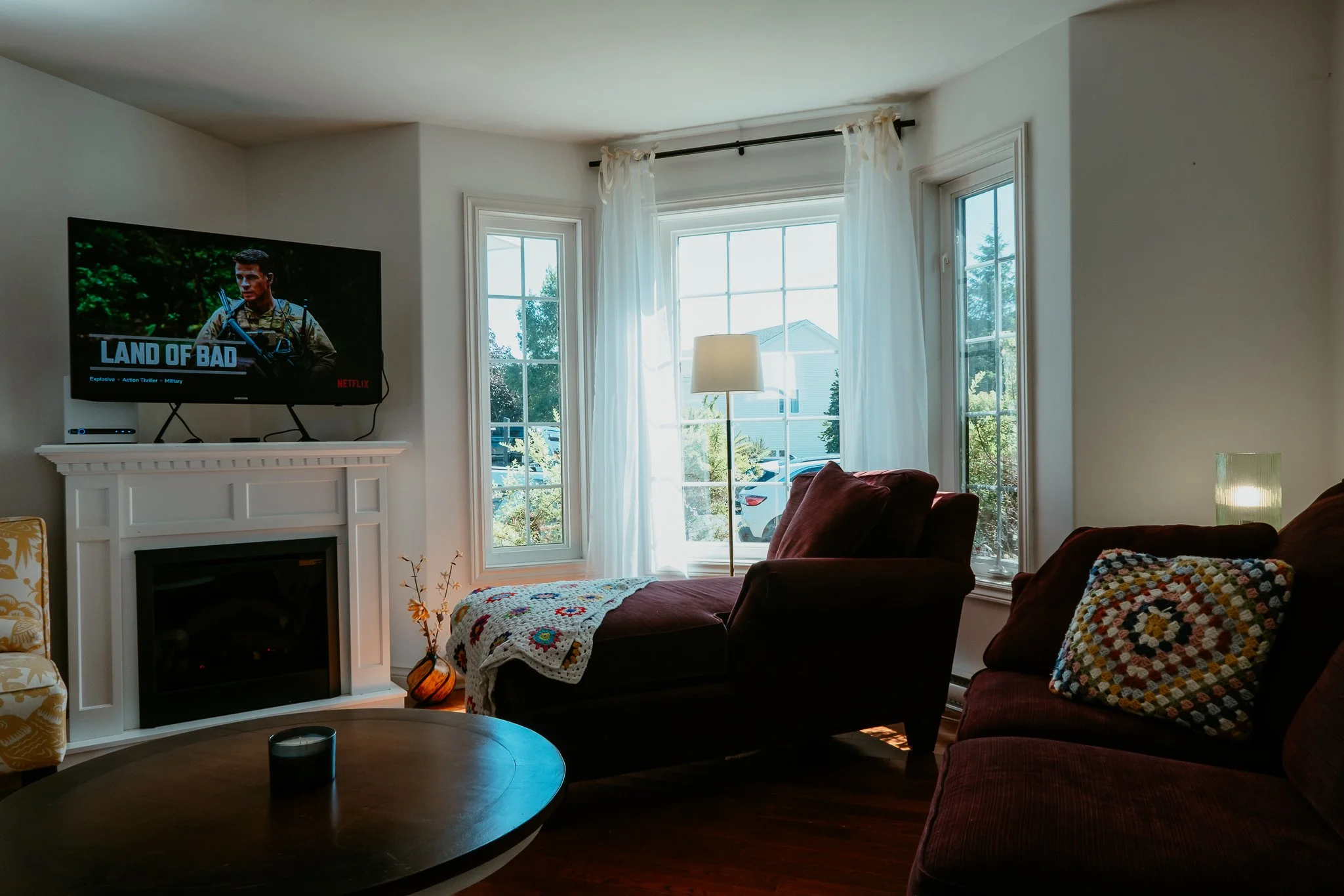 Living room with a fireplace, a flat-screen TV displaying 'Land of Bad' on Netflix, two red couches with crocheted pillows, a round wooden coffee table, a floor lamp, and three large windows with white curtains, allowing sunlight to brighten the space.