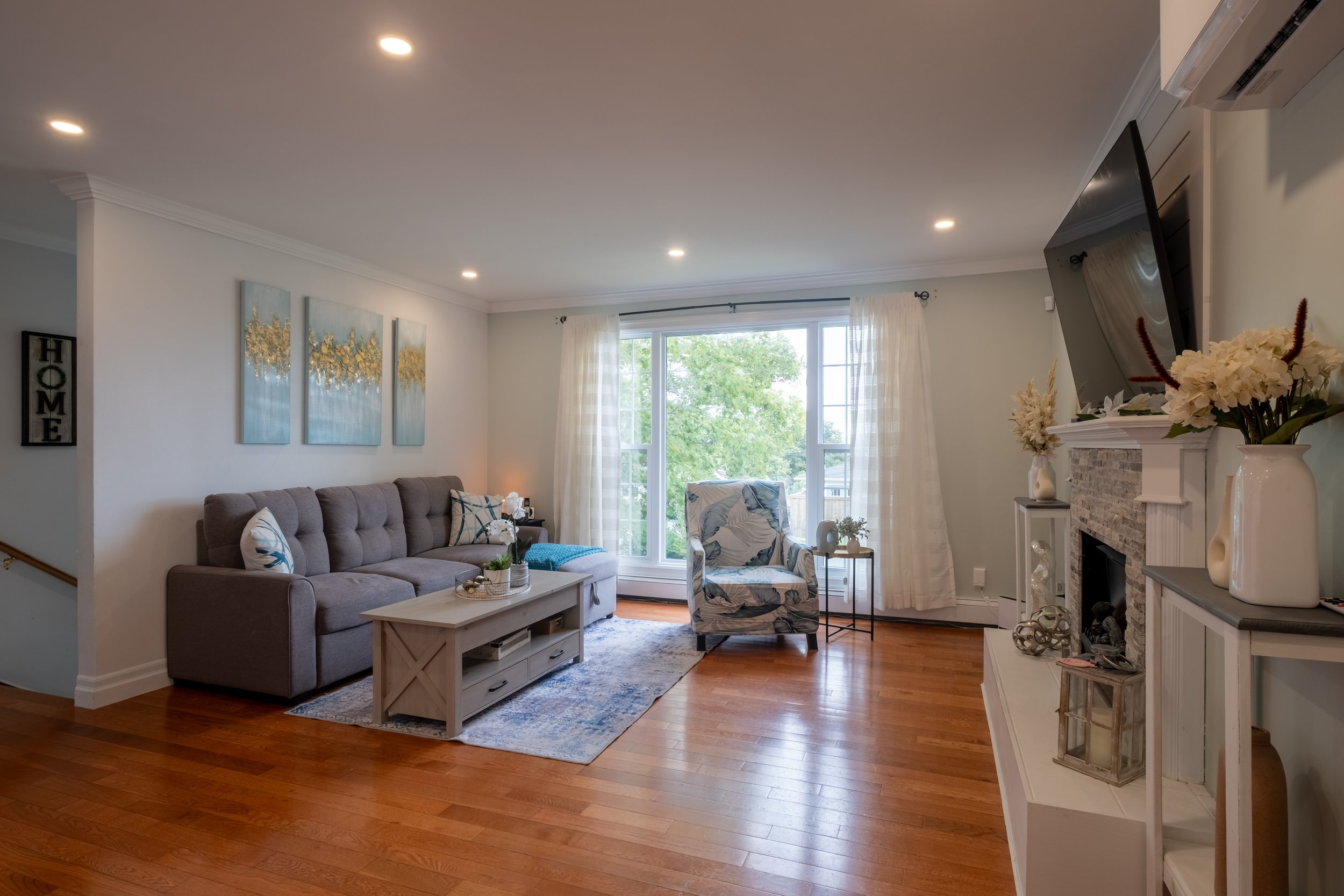 Living room with hardwood floors, a gray sofa with pillows, a coffee table, a patterned armchair, a large window with white curtains, wall art, and a fireplace decorated with flowers and vases.