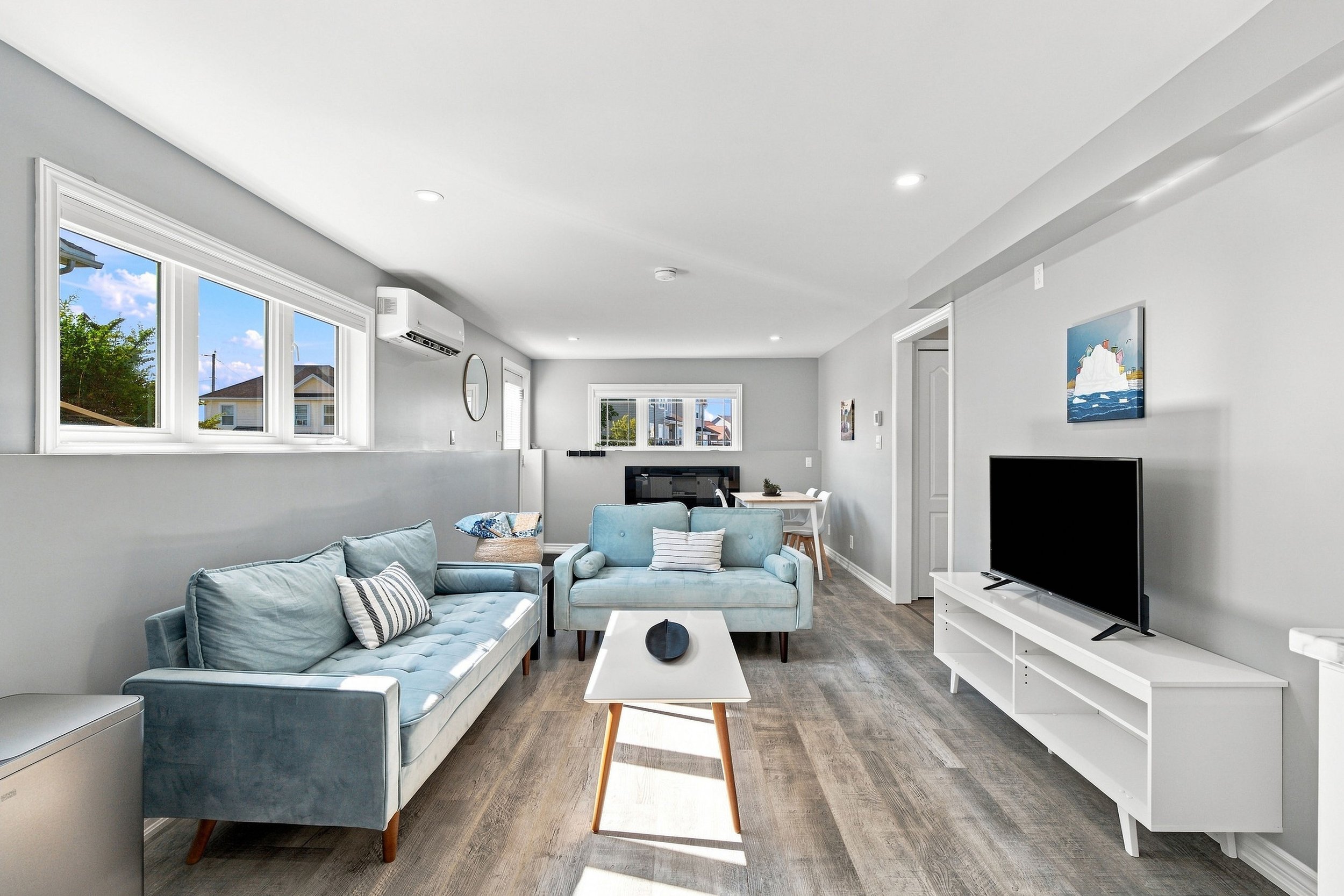 Living room with light gray walls, white ceiling, three large windows showing a suburban neighborhood, and a wall-mounted air conditioning unit. Furniture includes a light blue tufted sofa with striped pillows, a white coffee table, a small round side table, a dining table with chairs, a TV on a white stand, and wall art of an iceberg. The room has wood flooring and modern recessed lighting.