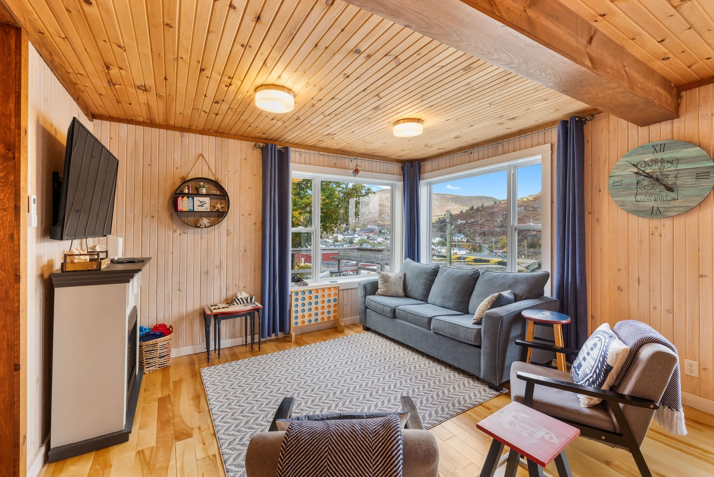A cozy living room with wooden walls and ceiling, featuring large windows with blue curtains, a gray sofa, a patterned area rug, a wall clock, a small side table, and a view of hills and houses outside.