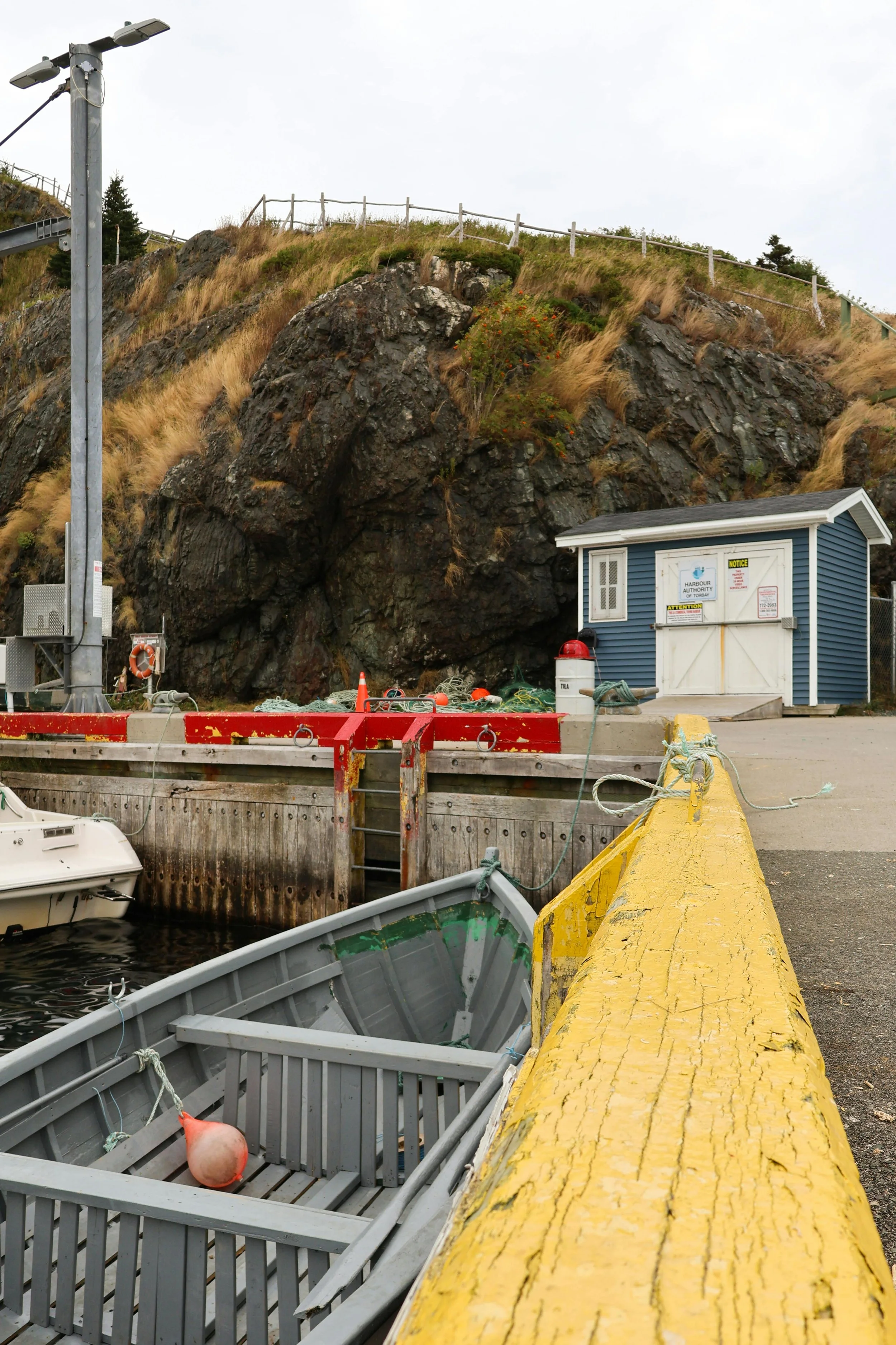 View of a harbor dock with boats, a yellow pier, a blue shed, a rocky hillside, and a streetlight.