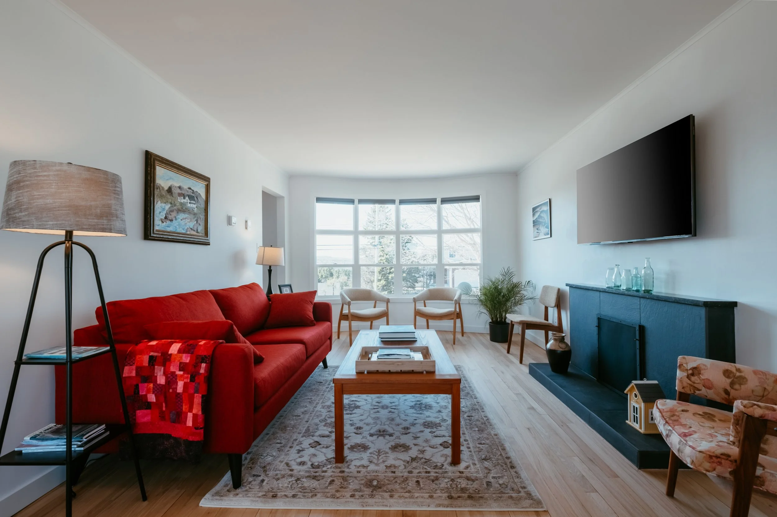 Living room with red sofa, beige chairs, wooden table, large window, wall-mounted TV, artwork, and decorative items.