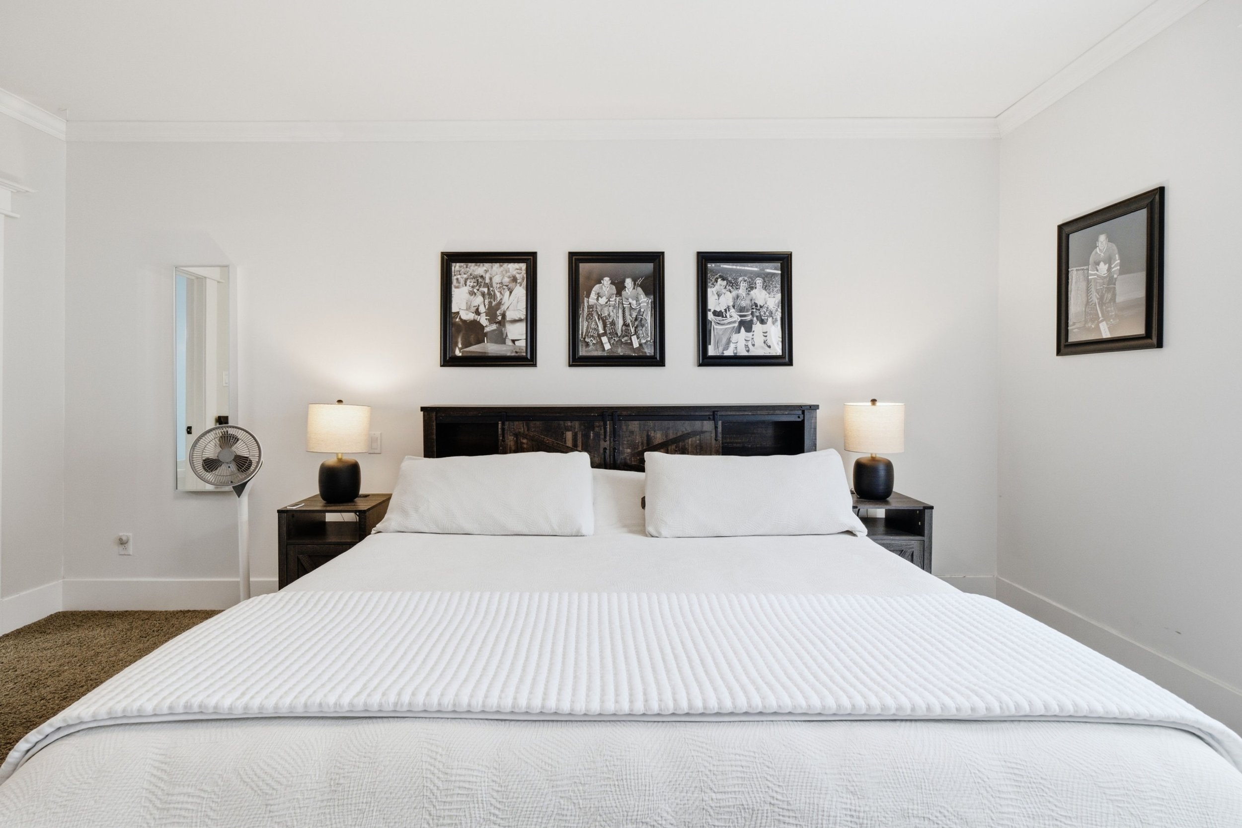 A bedroom featuring a neatly made bed with white bedding, two lamps on dark nightstands, black-and-white framed photographs on the wall above the bed, a fan on the left side, and cream carpeted flooring.